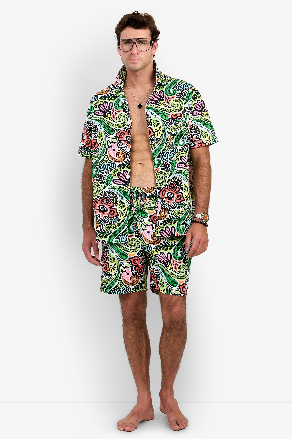 A man stands barefoot against a plain white background, wearing the Short Sleeve Boxer Pajamas Set featuring a matching colorful paisley pattern. The shirt is unbuttoned to reveal his chest, and he accessorizes with glasses, necklaces, a watch, and has light brown hair.