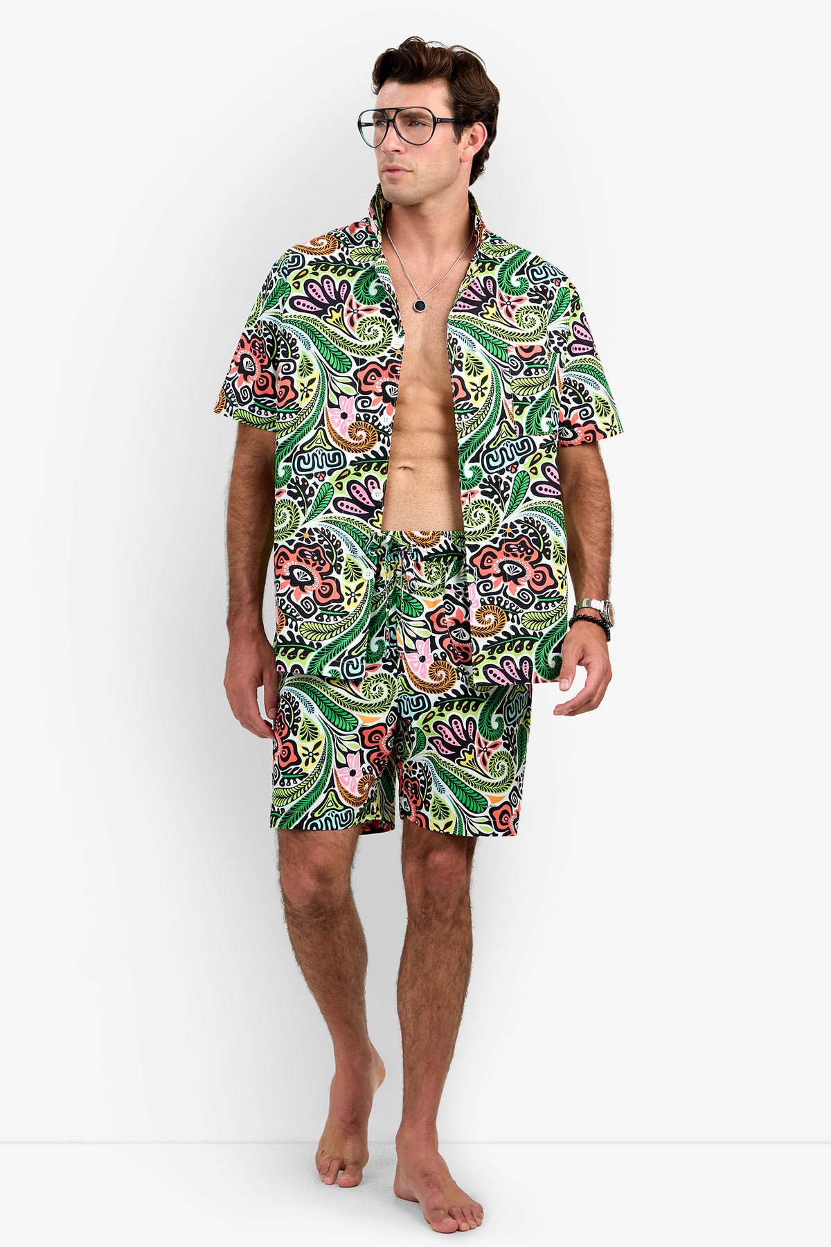A man wearing glasses stands barefoot against a white background, modeling the Short Sleeve Boxer Pajamas Set with the shirt open to reveal his chest.
