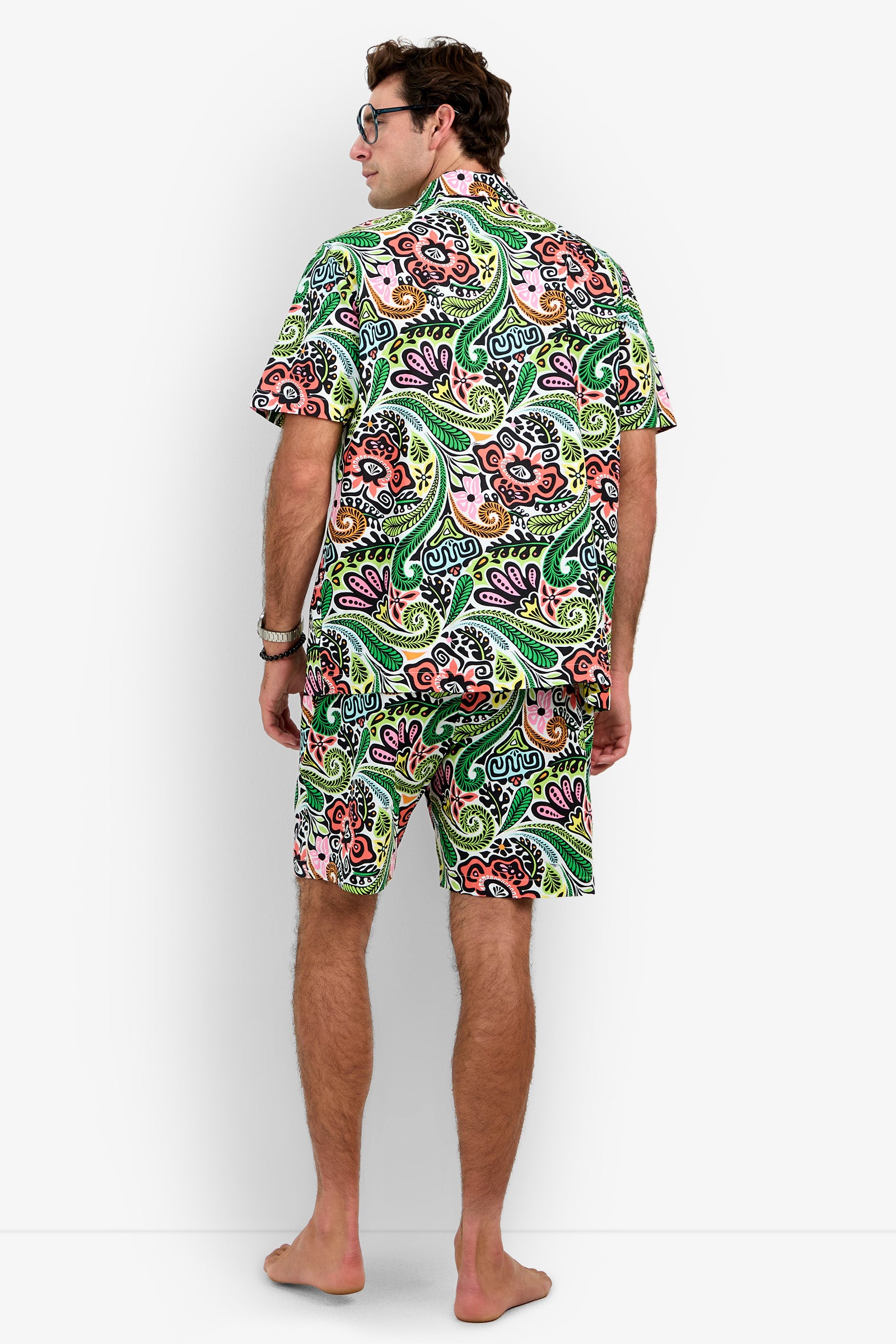 A barefoot man stands facing away, wearing the Short Sleeve Boxer Pajamas Set featuring a matching green, pink, and orange paisley pattern. He has short brown hair and glasses, with his hands relaxed by his sides.