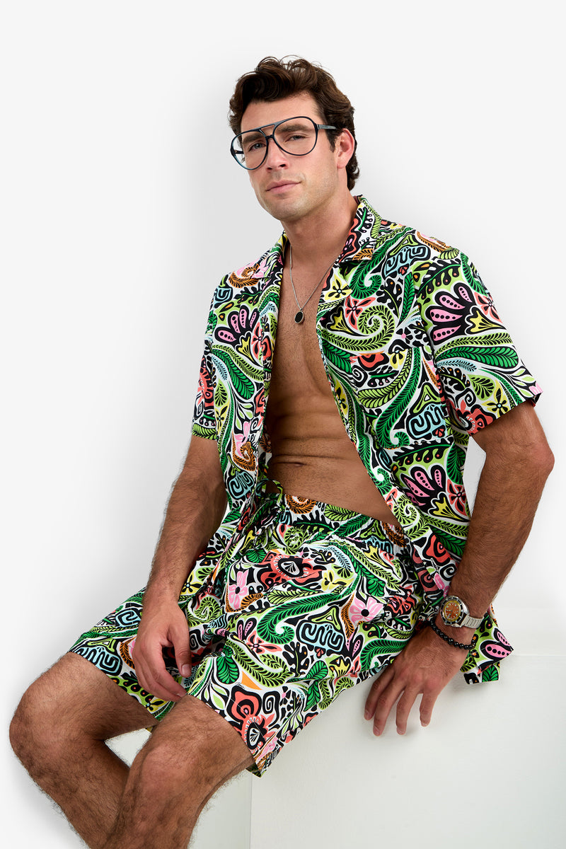 A man with glasses wears the Short Sleeve Boxer Pajamas Set, featuring a vibrant, tropical pattern. He sits on a white surface with the shirt open to reveal his chest, exuding a relaxed and confident vibe.