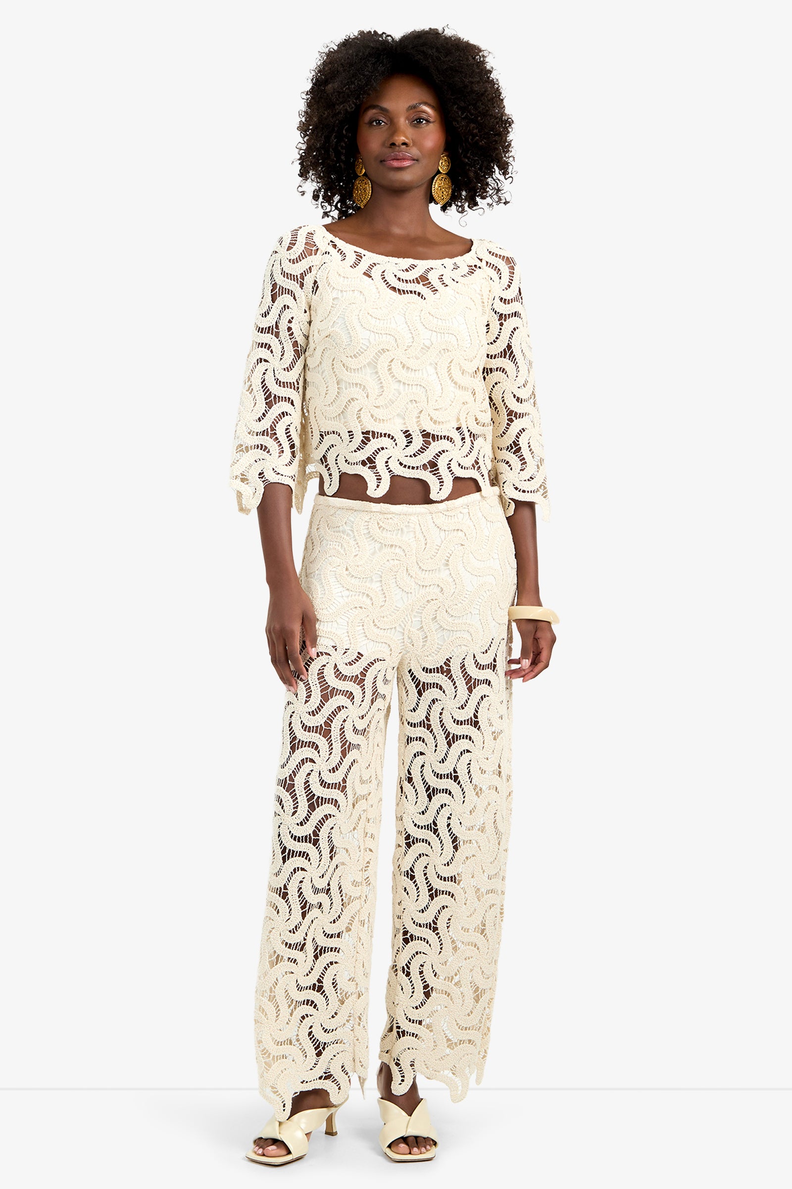 A woman with short curly hair, gold earrings, and heeled sandals stands facing forward in the Helen Top—a cream-colored, see-through lace top with a wavy pattern—paired with matching wide-leg pants.