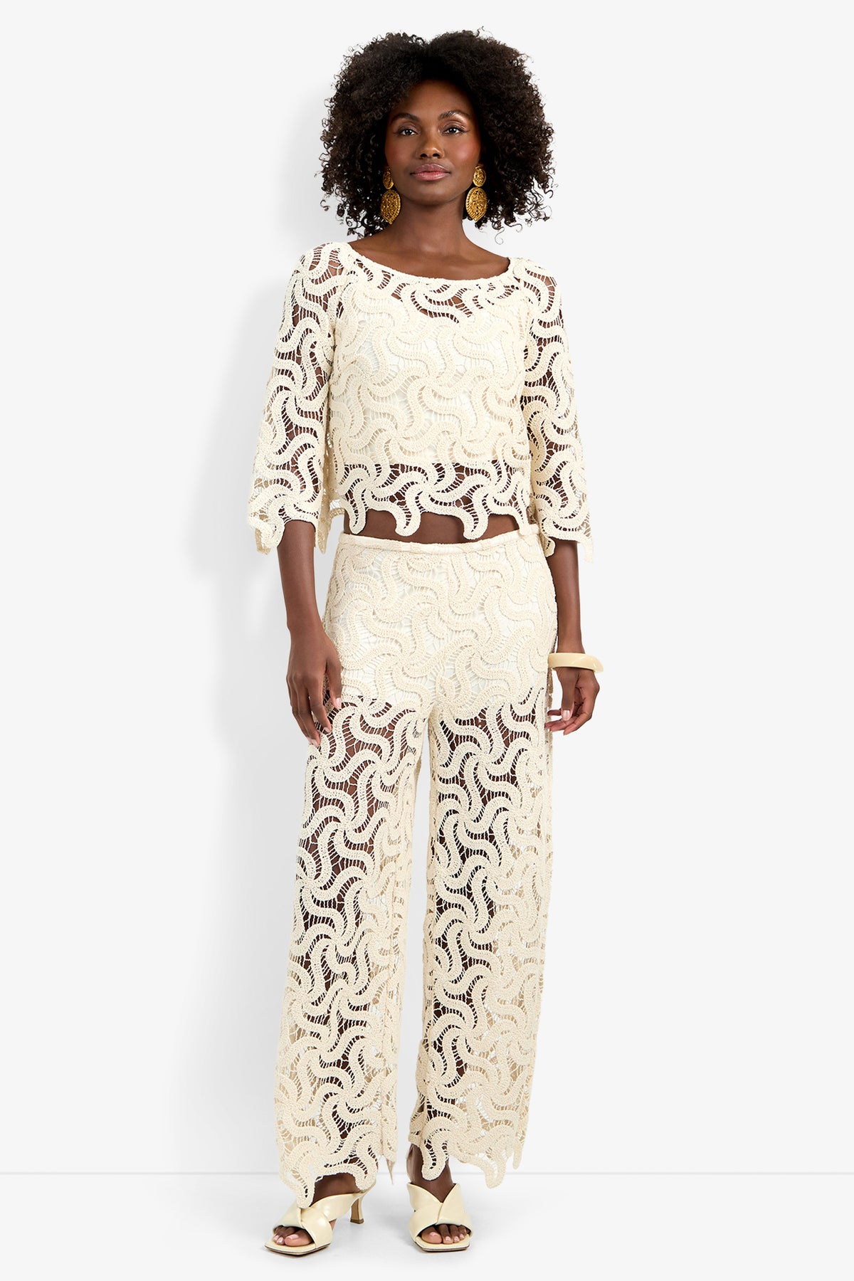 A woman with curly hair and statement earrings wears the Helen Top—a cream, lacy, semi-sheer blouse—with wide-leg wavy pants and heeled sandals, standing against a white background.
