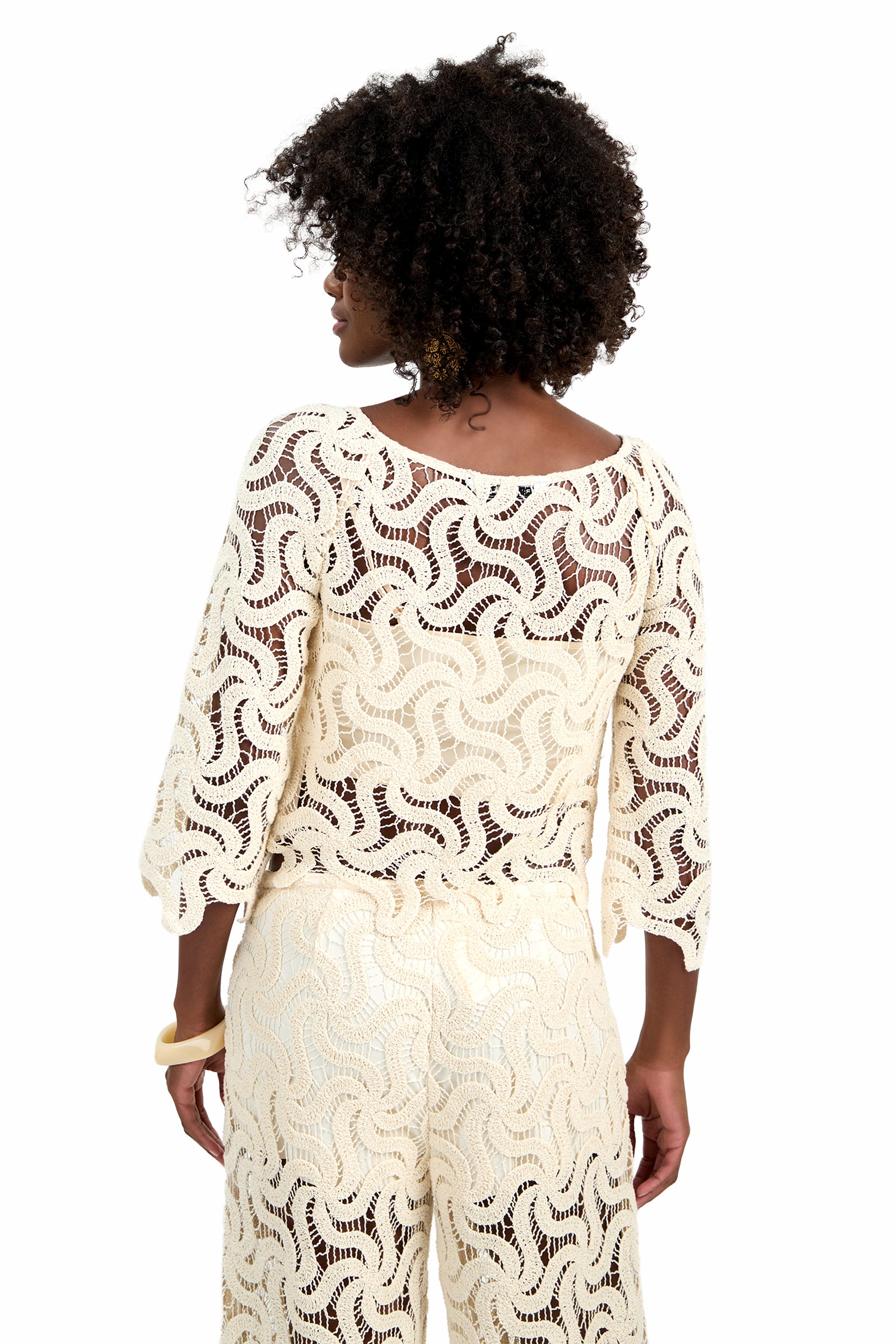 Woman with curly hair faces away, wearing the Helen Top in cream—an open-knit crochet design paired with matching pants. The white background highlights the texture and style of the outfit.