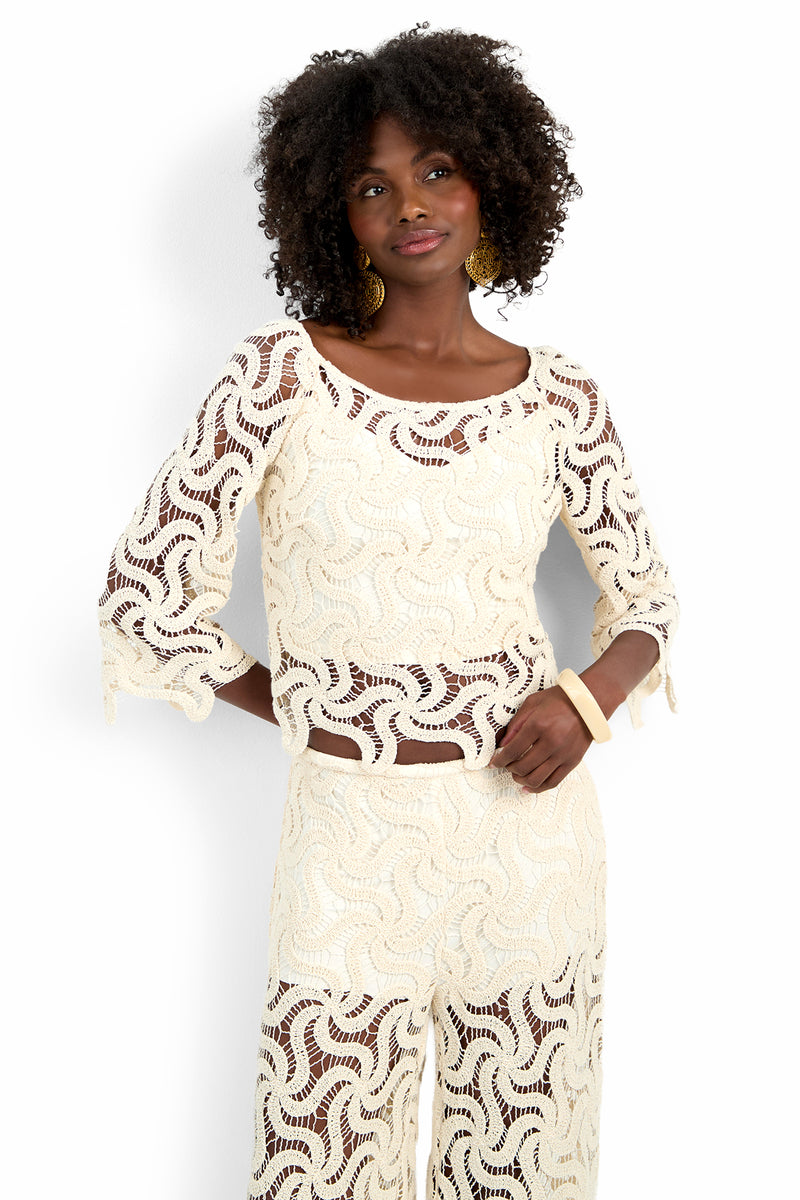 A woman with curly hair wears the matching cream-colored, crocheted Helen Top and pants with a wavy pattern, accessorized with gold hoop earrings and a chunky beige bracelet, standing against a white background.