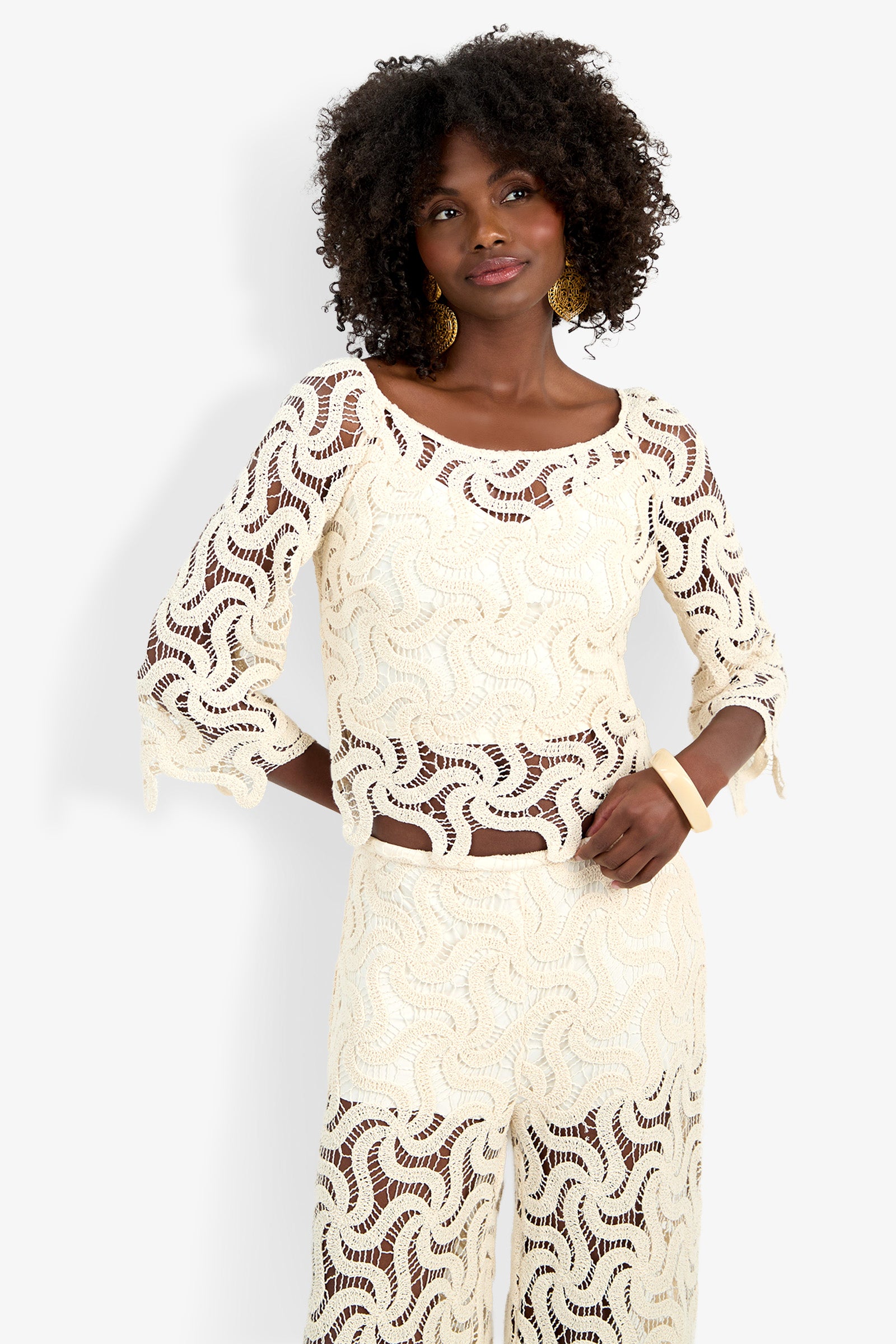A woman with curly hair wears the Helen Top, a sheer, cream crochet style with a wavy pattern and matching pants. She accessorizes with gold earrings and a white bracelet, standing hands on hips against a white background.