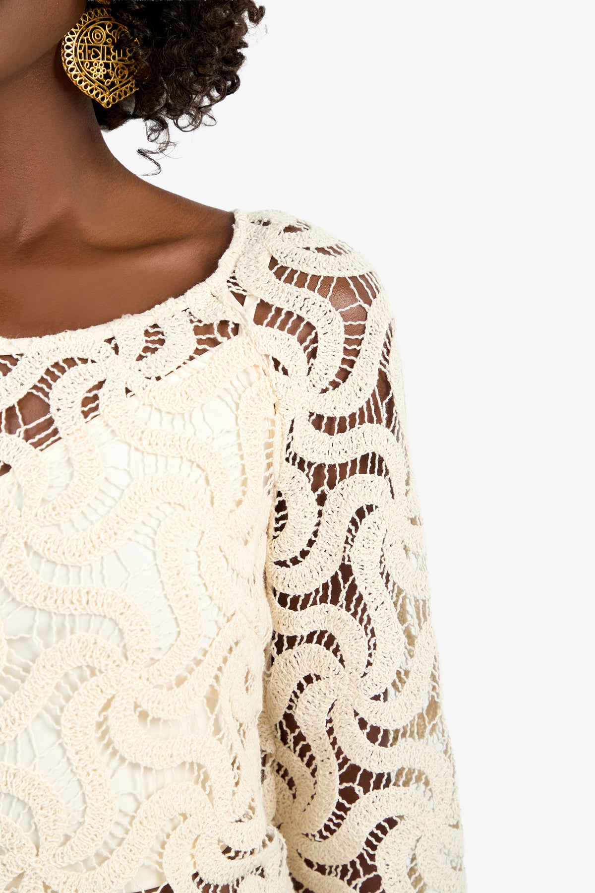 A close-up of a woman wearing the Helen Top, a cream-colored, intricately crocheted piece with wavy patterns, paired with a large round gold earring. Her face is not visible.