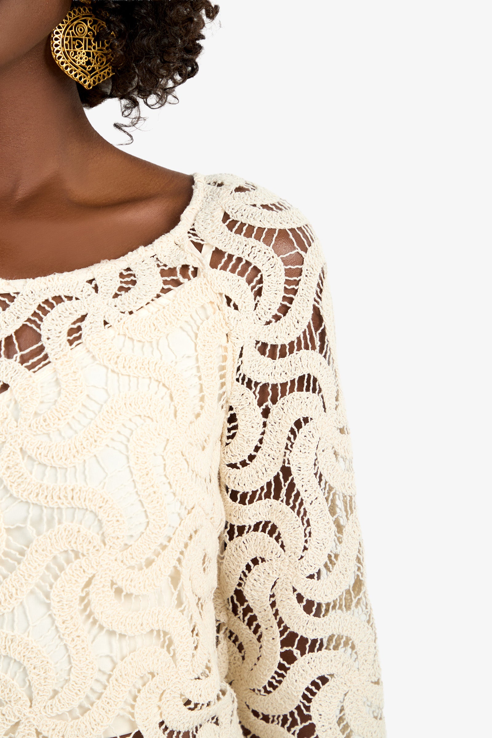 A close-up of a woman wearing the Helen Top, a cream-colored, intricately crocheted piece with wavy patterns, paired with a large round gold earring. Her face is not visible.
