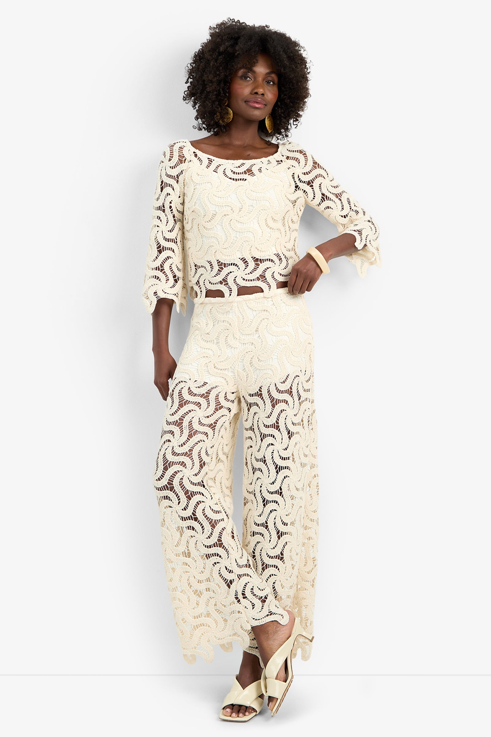 A woman with curly hair poses with one hand on her hip, wearing the cream-colored, semi-sheer Helen Top and matching wide-leg pants, styled with gold earrings, a bracelet, and cream heeled sandals.