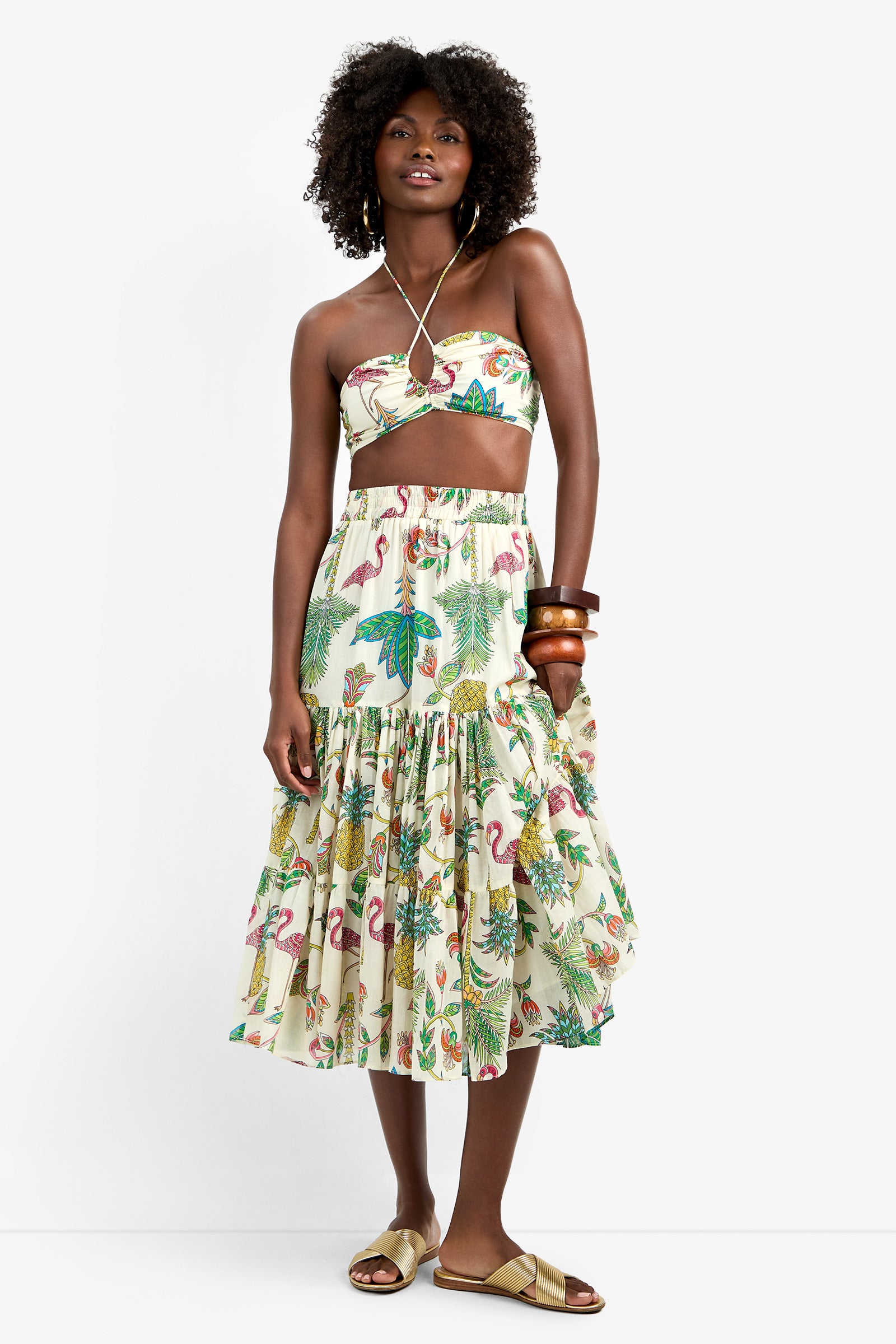 A woman wears the Marisalle Top, a tropical print halter crop top, paired with a flared midi skirt. She accessorizes with large wooden bangles and gold sandals, standing against a plain white background.