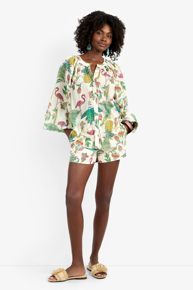 A woman wears the NORRY TOP, a loose long-sleeved button-down shirt in a colorful tropical print made of soft 100% cotton, paired with matching shorts and beige slide sandals, with one hand in her pocket.