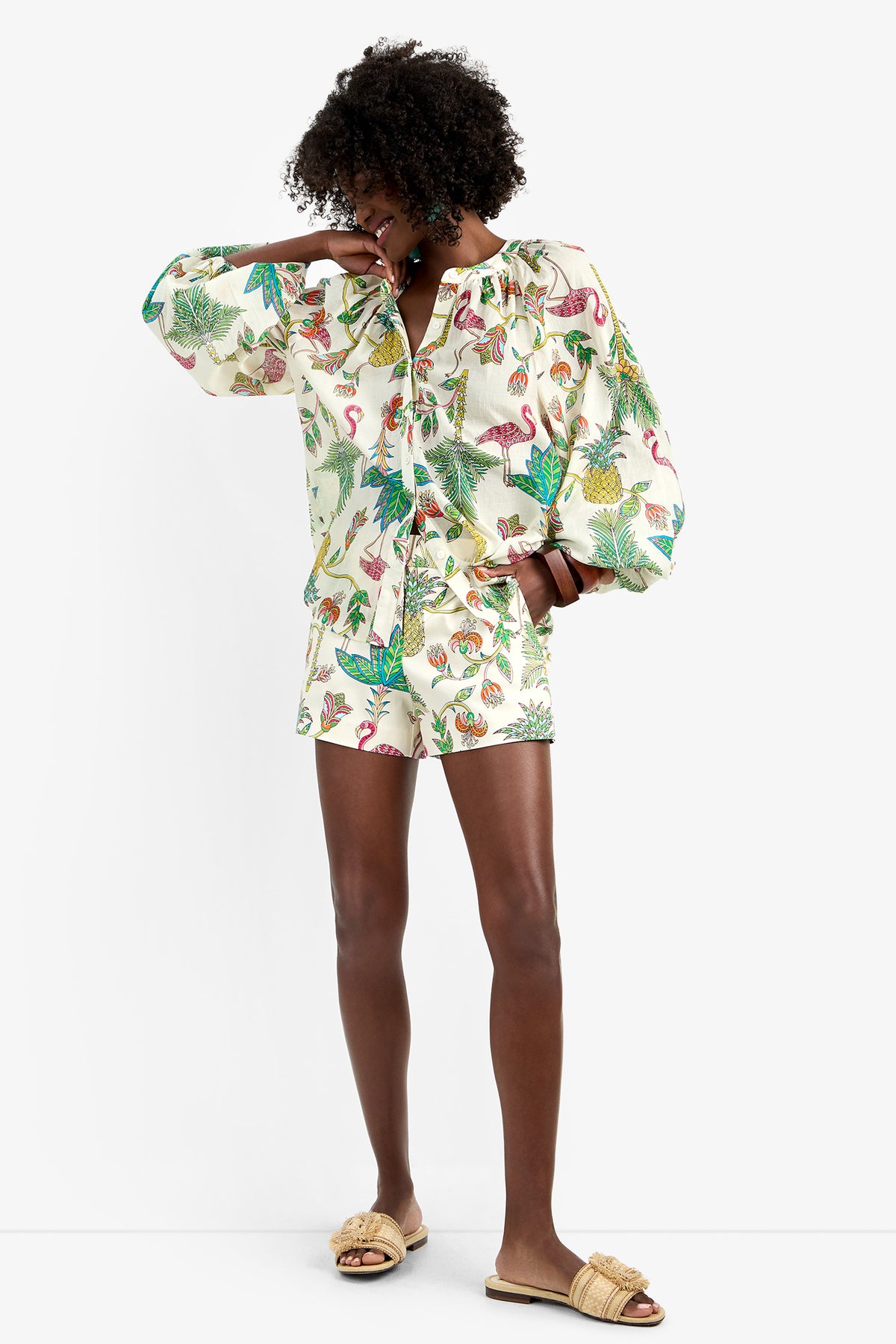 A person smiles in the NORRY TOP, a matching set with tropical flamingo and leaf prints made from 100% cotton. They pair it with tan slide sandals, raising one arm to partially cover their face against a plain white background.