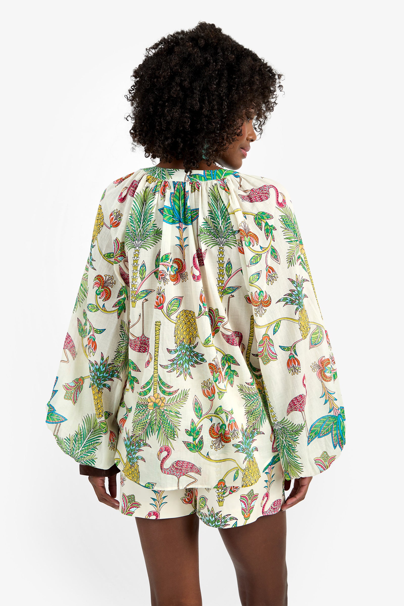 A person with curly hair, seen from behind, wears the NORRY TOP—100% cotton, long-sleeved and loose—with a colorful tropical print of plants, birds, and fruit on a white background.