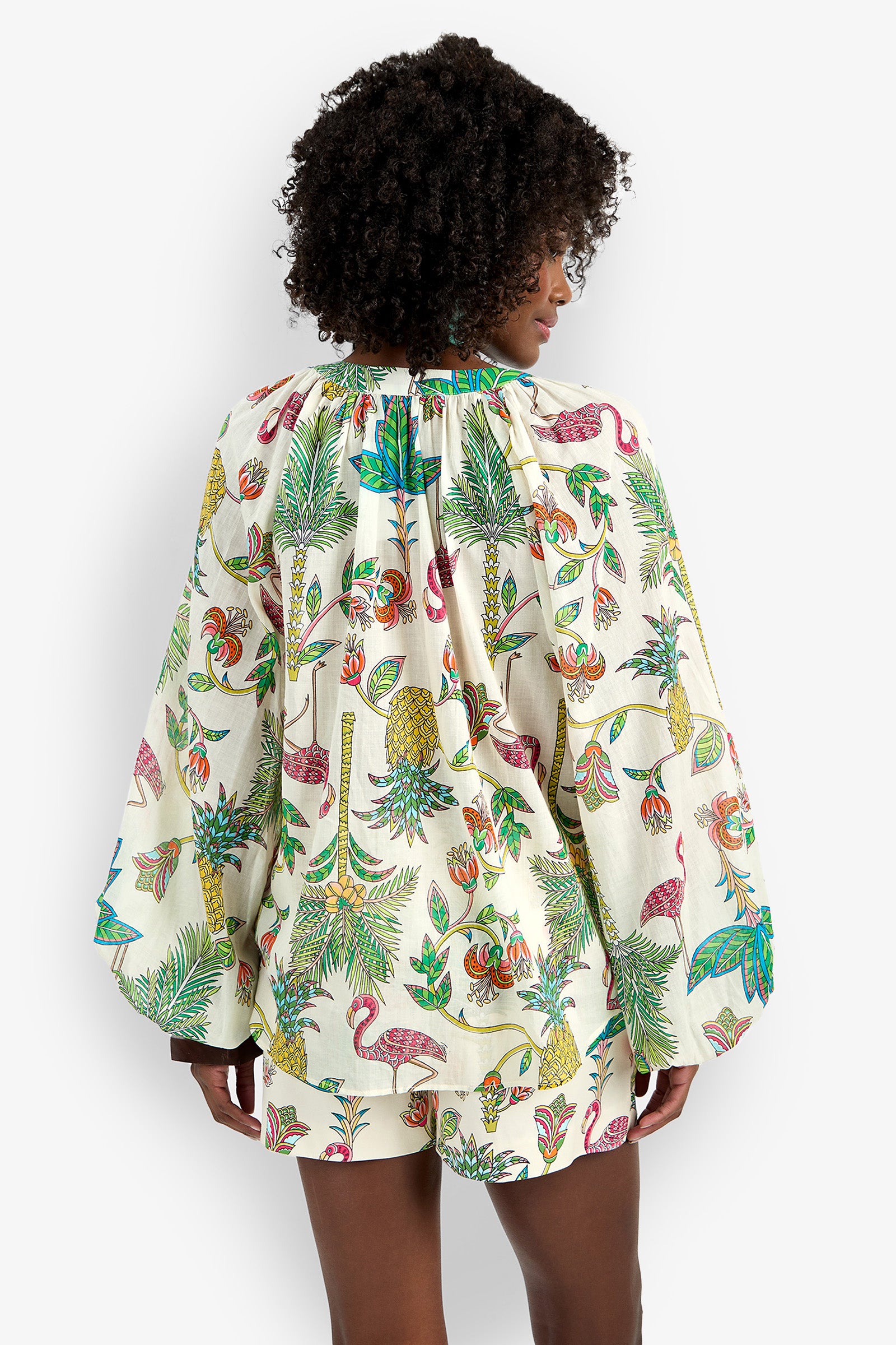 A woman with curly hair faces away, wearing the Norry Top and matching shorts, both featuring a vibrant tropical print with flamingos, pineapples, flowers, and leaves on a cream background.