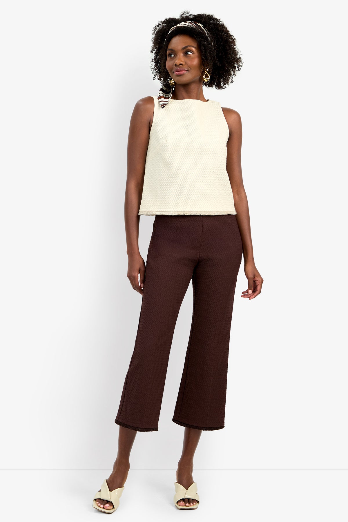 A woman stands against a white background wearing the sleeveless cream Port Top, brown textured cropped pants, cream open-toe heels, gold earrings, and a patterned headband.