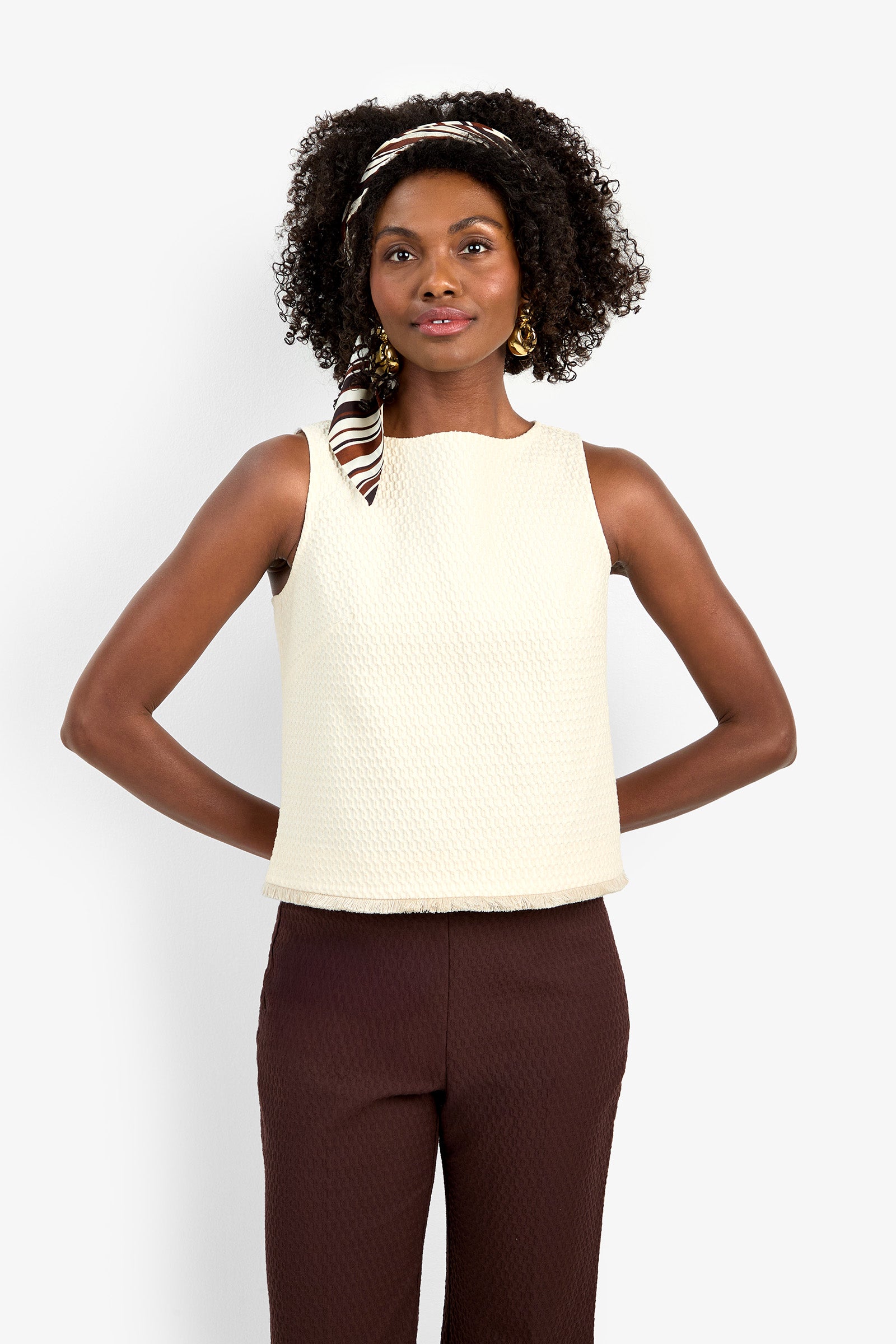 A woman with curly hair, wearing the Port Top and brown pants, stands with her hands behind her back against a plain white background.