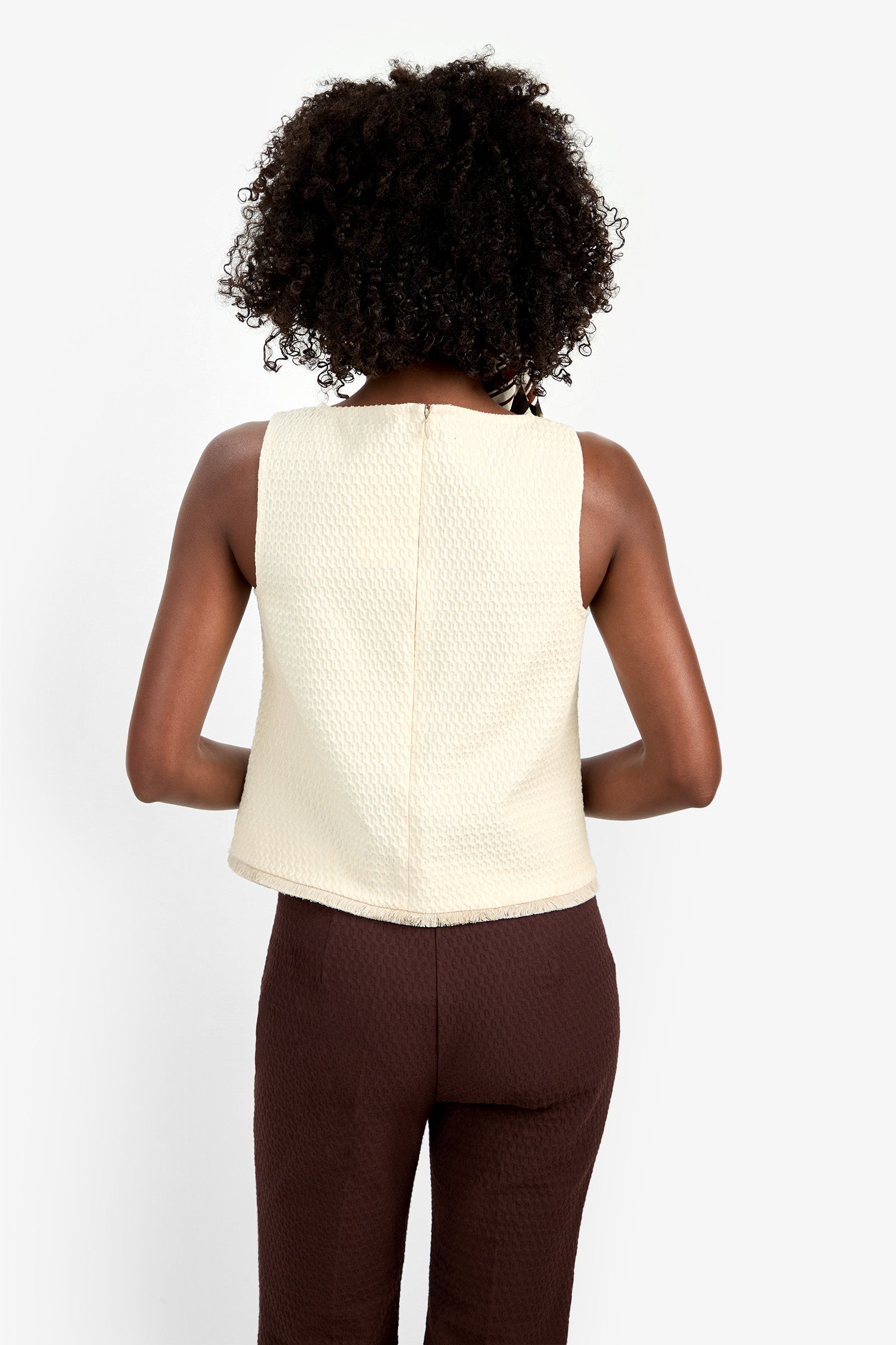 A person with curly hair is shown from the back, wearing the sleeveless Port Top in beige and high-waisted brown pants against a plain white background.