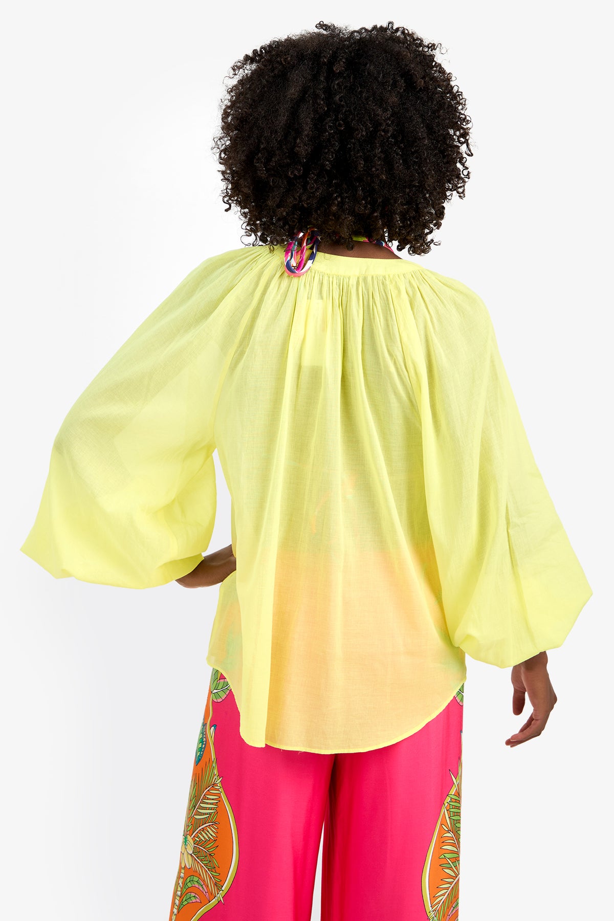 A person with curly hair is seen from behind, wearing the NORRY TOP—a loose, light yellow, 100% cotton long-sleeve blouse—paired with bright pink pants featuring colorful patterns against a plain white background.