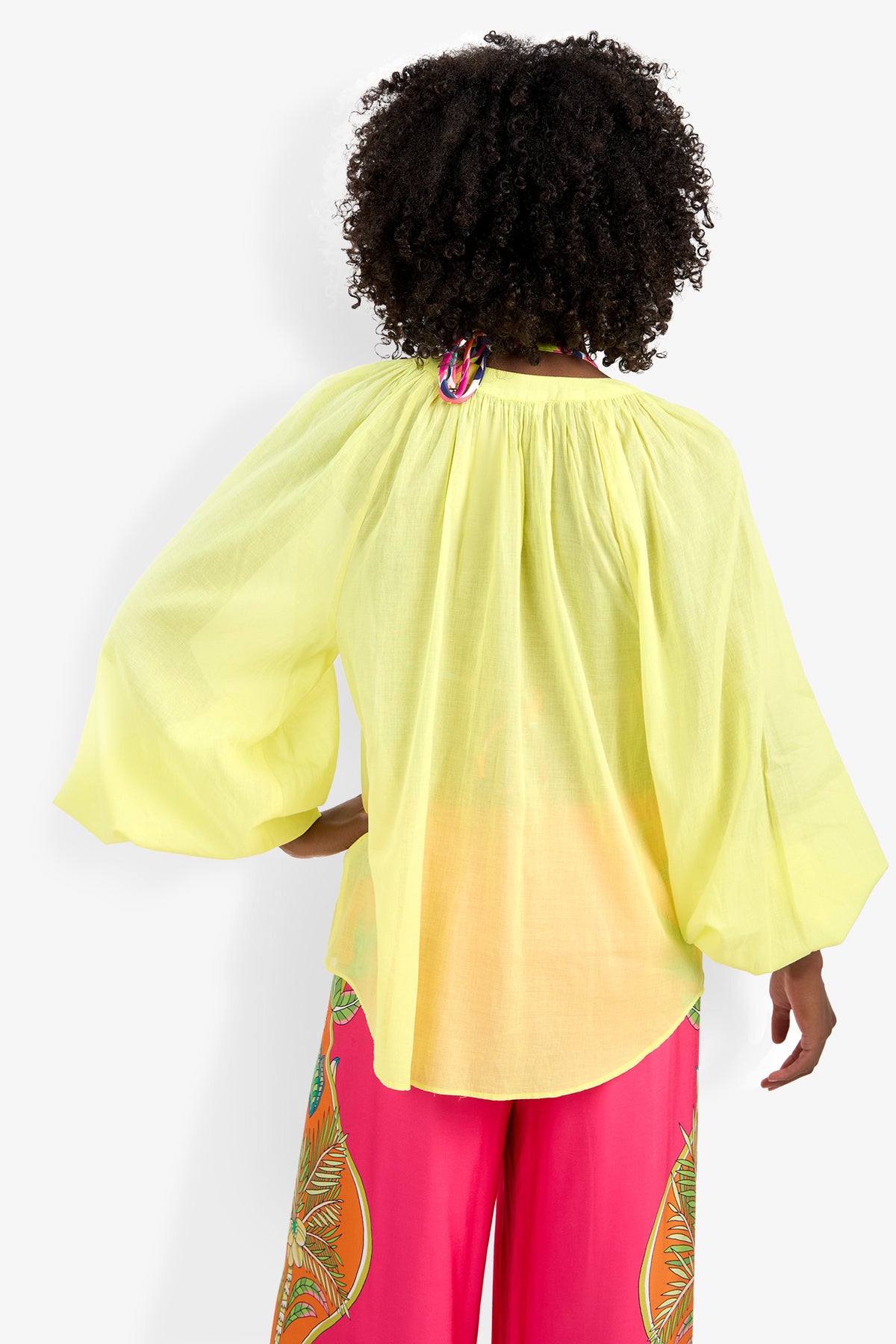 A person with curly hair, seen from behind, wears the Norry Top—a loose, yellow ombré blouse with wide sleeves—paired with bright pink patterned pants for a vibrant, summery look.