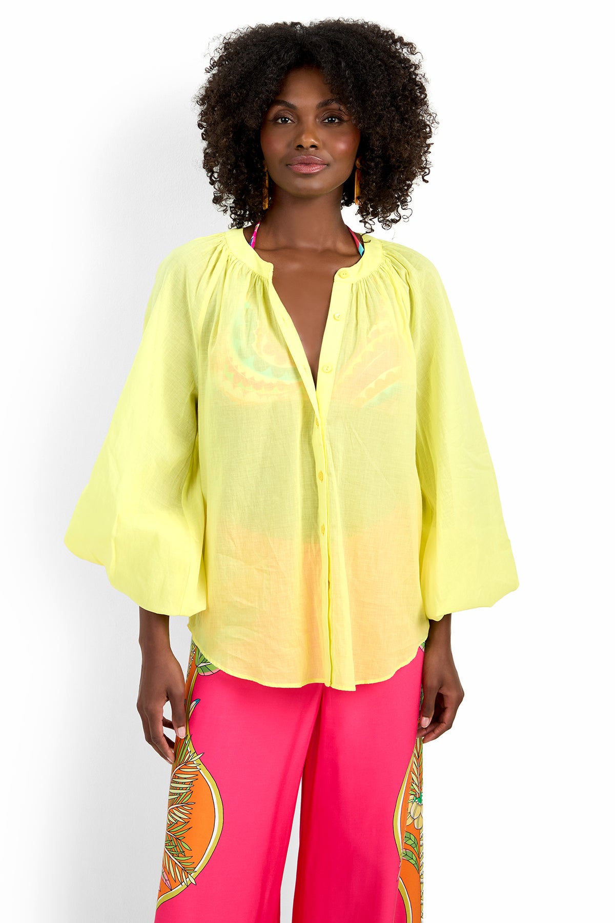 A woman models the NORRY TOP, a loose, light yellow 100% cotton button-up blouse with wide sleeves, paired with bright pink pants with a colorful side pattern, standing against a plain white background.