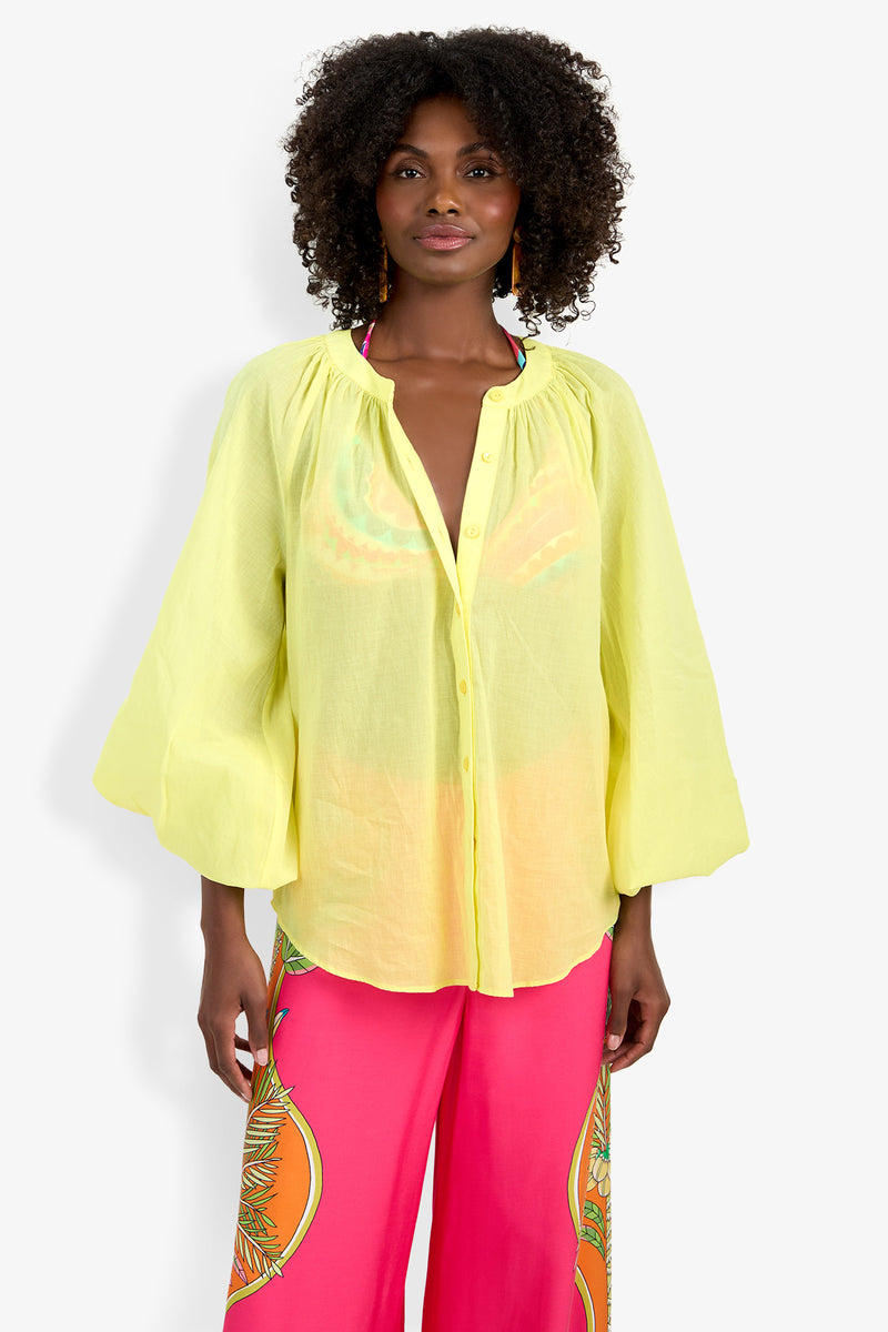 A woman wears the Norry Top, a sheer, loose-fitting yellow button-up blouse with wide sleeves, paired with bright pink patterned pants, standing against a plain white background.