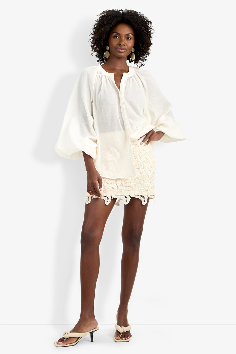 A woman wearing the Norry Top—a loose, long-sleeved white blouse—paired with a cream mini skirt, dangling earrings, and heeled sandals stands confidently against a plain white background.