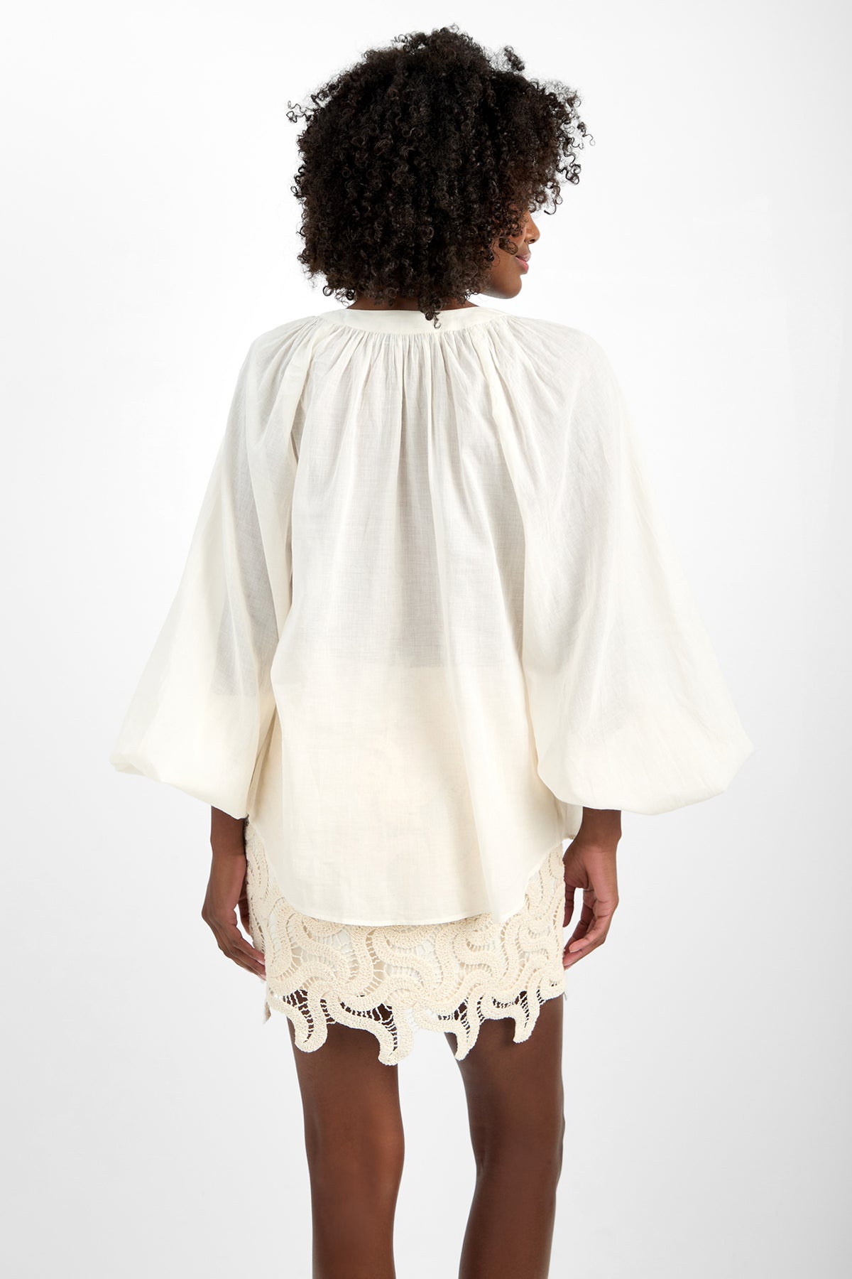 A woman with curly hair stands facing away, wearing the loose, white 100% cotton NORRY TOP long-sleeve blouse and a cream crochet mini skirt with a wavy hem against a plain white background.