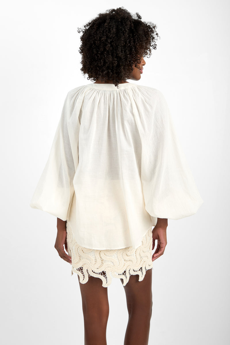 A woman with curly hair stands facing away, wearing the loose, white 100% cotton NORRY TOP long-sleeve blouse and a cream crochet mini skirt with a wavy hem against a plain white background.
