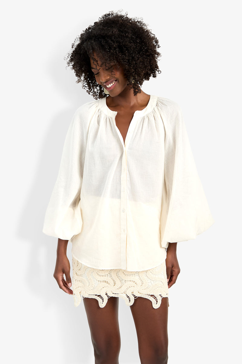 A smiling woman with curly hair wears the Norry Top, a loose, long-sleeved off-white button-up blouse. She pairs it with a cream crochet mini skirt and stands against a plain white background.