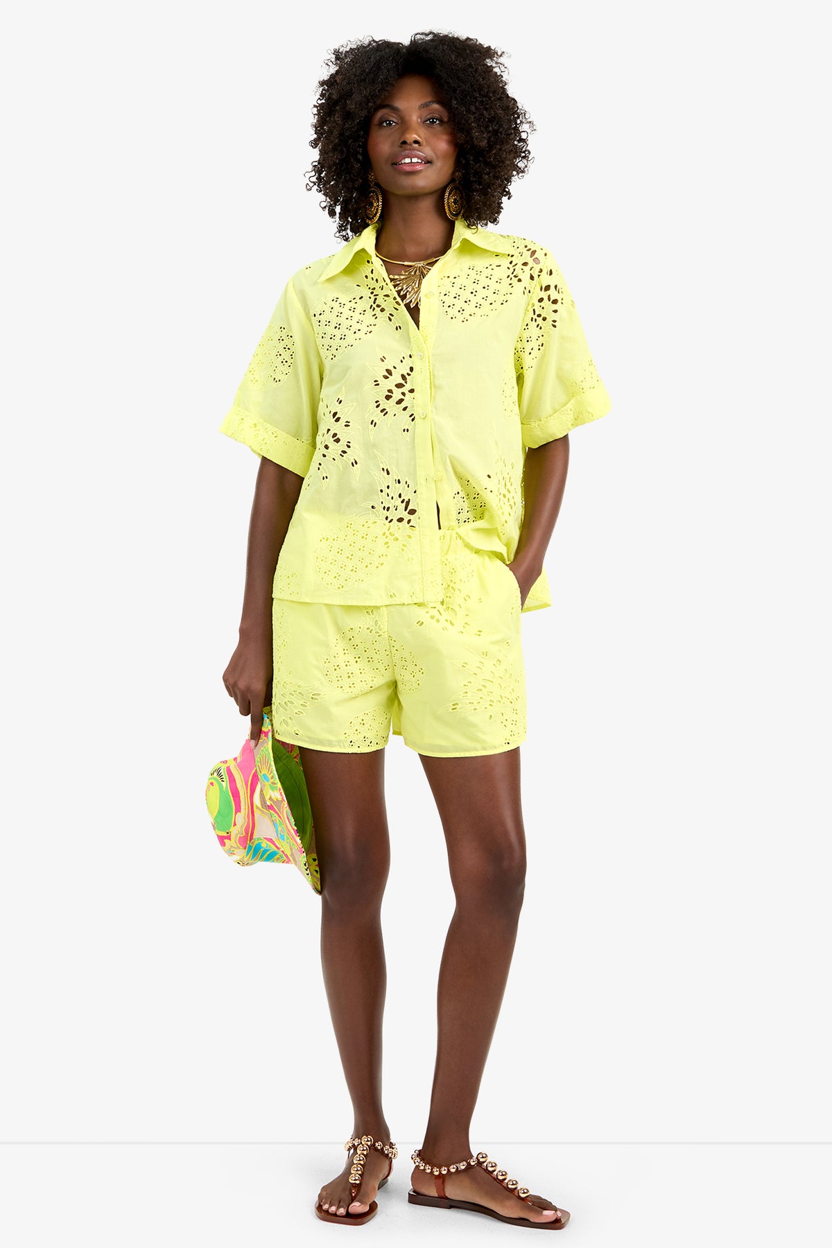 A woman stands against a white background wearing the TRIENNALE 2 TOP, a bright yellow 100% cotton eyelet short-sleeve button-up shirt, with matching shorts, colorful clutch, hoop earrings, layered necklaces, and sandals.