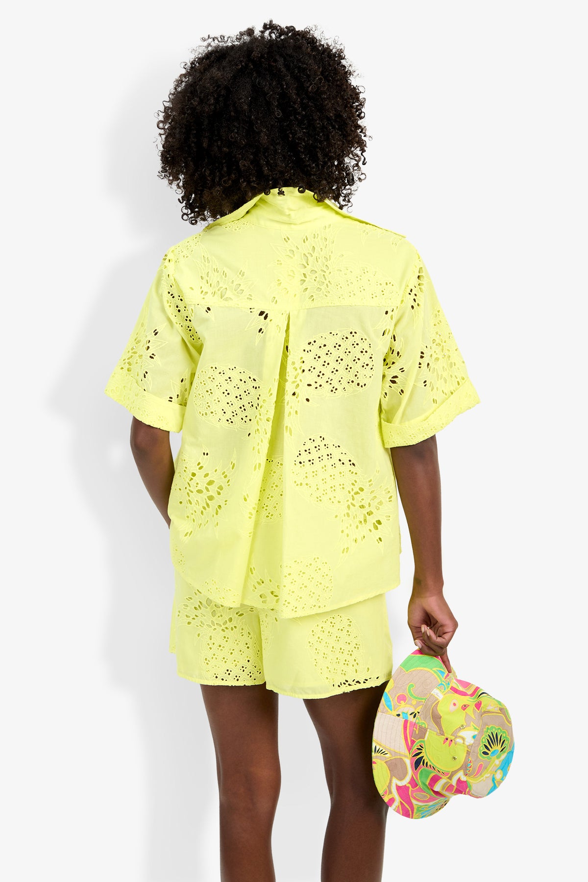 A person with curly hair, seen from behind, wears the bright yellow Triennale Top and matching shorts, holding a colorful patterned hat in their left hand.