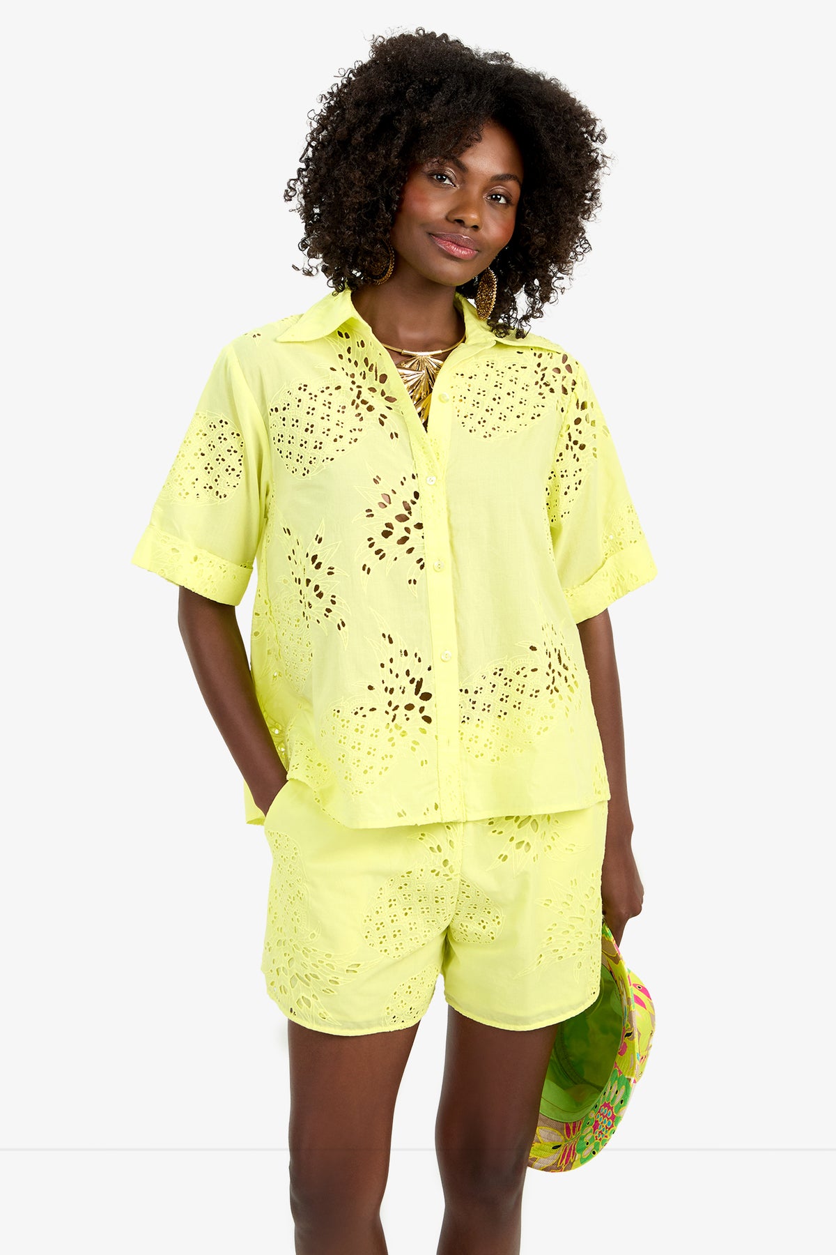 A woman wears the TRIENNALE 2 TOP, a light yellow, short-sleeve 100% cotton button-up shirt with laser-cut eyelet details and matching shorts, holding a colorful hat and smiling at the camera.