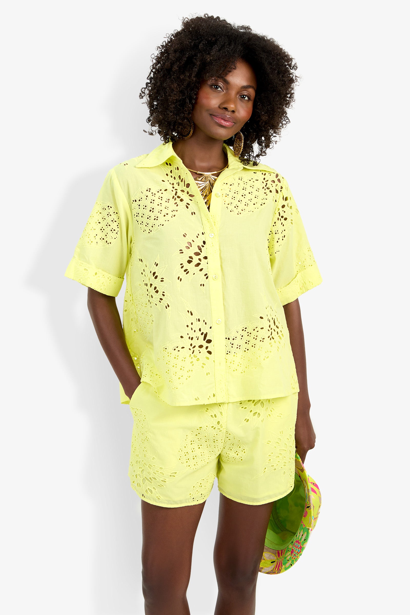 A woman wearing the Triennale Top, a yellow short-sleeve button-up shirt, with matching eyelet shorts stands against a white background, holding a colorful hat and smiling slightly.