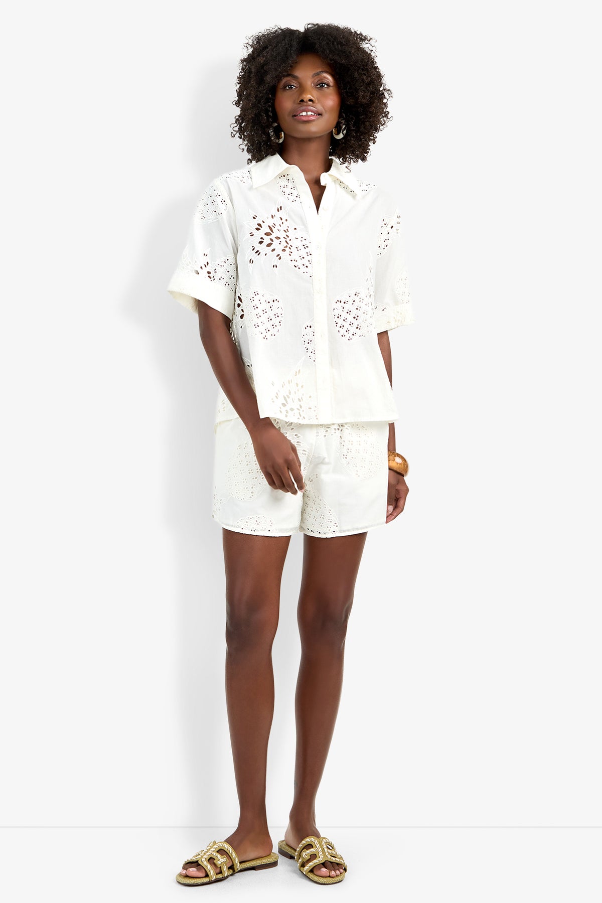 A woman wears the Triennale Top, a white short-sleeve button-up with eyelet details, paired with matching shorts. She has curly hair, gold earrings, and gold-toned sandals. Smiling, she stands with one hand in her pocket.