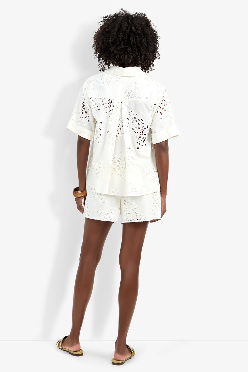 Someone with curly hair faces away, wearing the Triennale Top—a white, short-sleeve, eyelet button-up—paired with matching shorts, sandals, and a brown bracelet. The background is plain white.