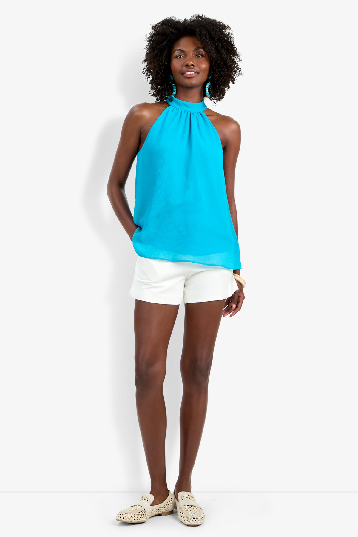 A woman with curly hair smiles slightly, hands in her pockets, wearing the sleeveless turquoise Kissimmee Top, white shorts, tan woven slip-ons, and large turquoise earrings against a white background.