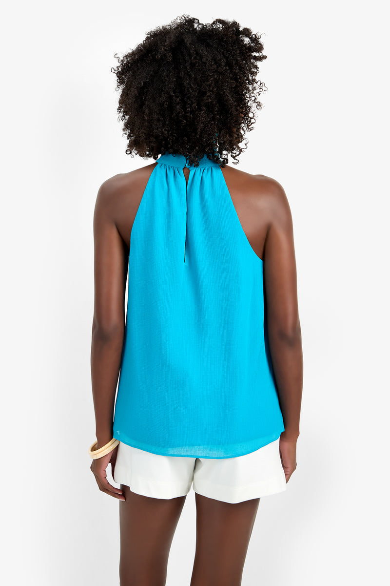 A person with curly hair is seen from behind wearing the KISSIMMEE TOP in bright blue (100% polyester) and white shorts, standing against a plain white background.