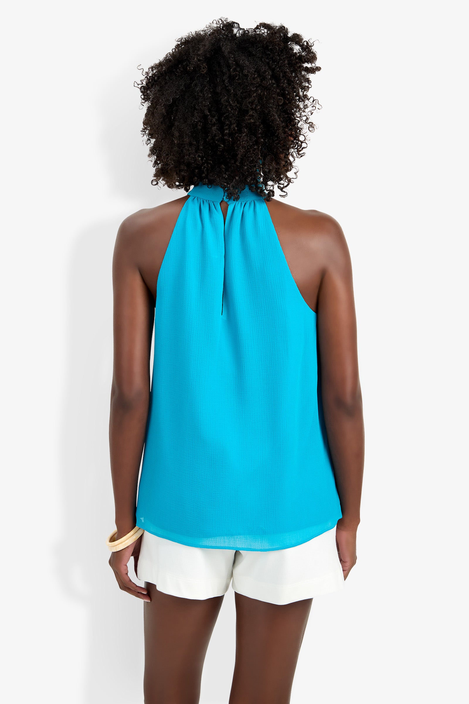 A person with curly hair, seen from behind, is wearing the bright blue Kissimmee Top and white shorts, accessorized with a bangle on their right wrist, all set against a plain white background.