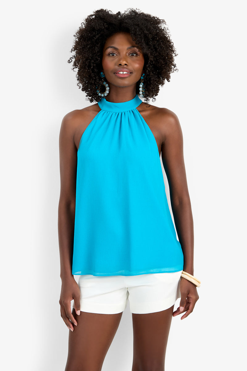 A woman wearing the KISSIMMEE TOP, a sleeveless bright blue halter made from polyester, pairs it with white shorts. She has curly hair, blue earrings, a bracelet, and stands smiling against a plain white background.