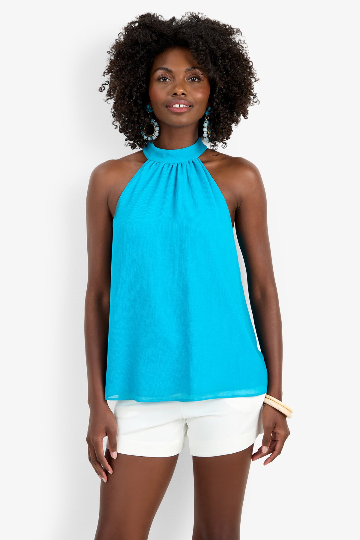 A woman wearing the KISSIMMEE TOP, a sleeveless bright blue halter made from polyester, pairs it with white shorts. She has curly hair, blue earrings, a bracelet, and stands smiling against a plain white background.