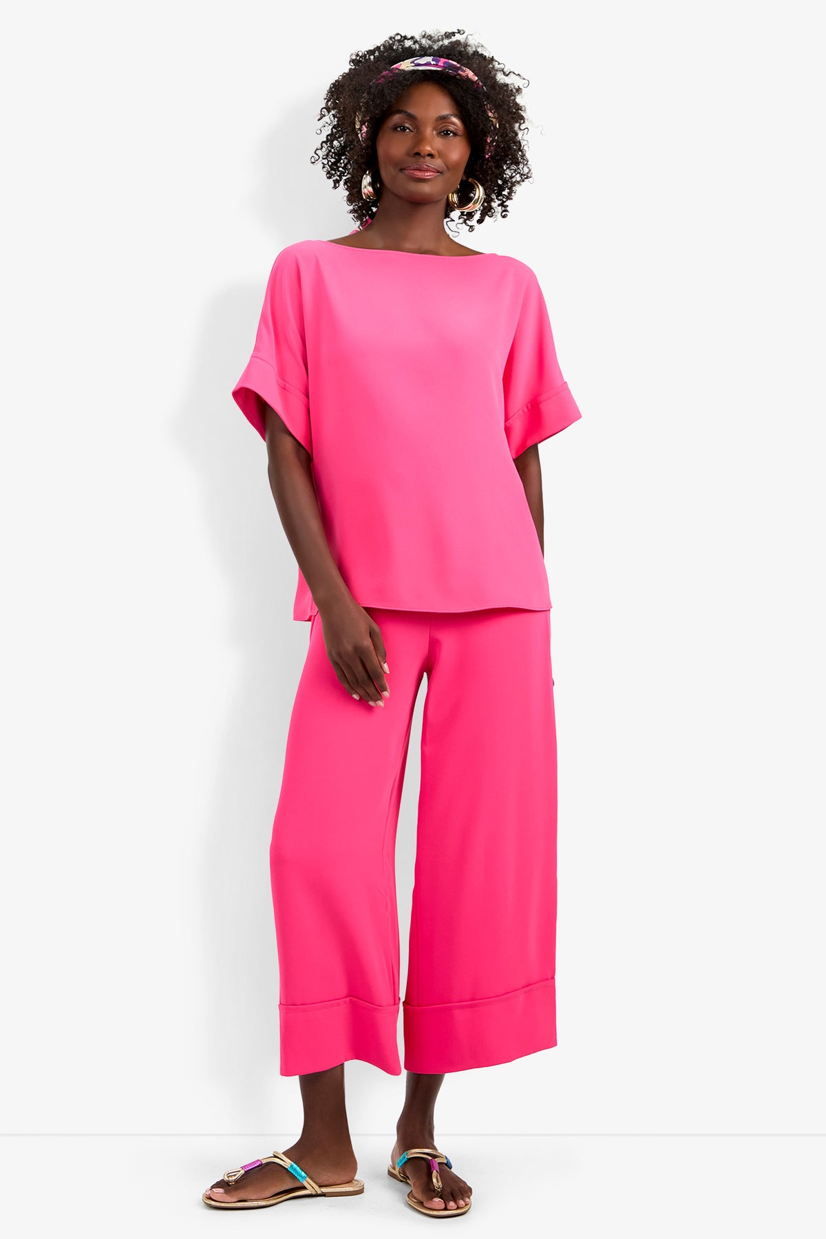A woman stands against a white background wearing the loose-fitting, bright pink Sofie Top and matching wide-leg pants. She has curly hair with a colorful headband and sandals.