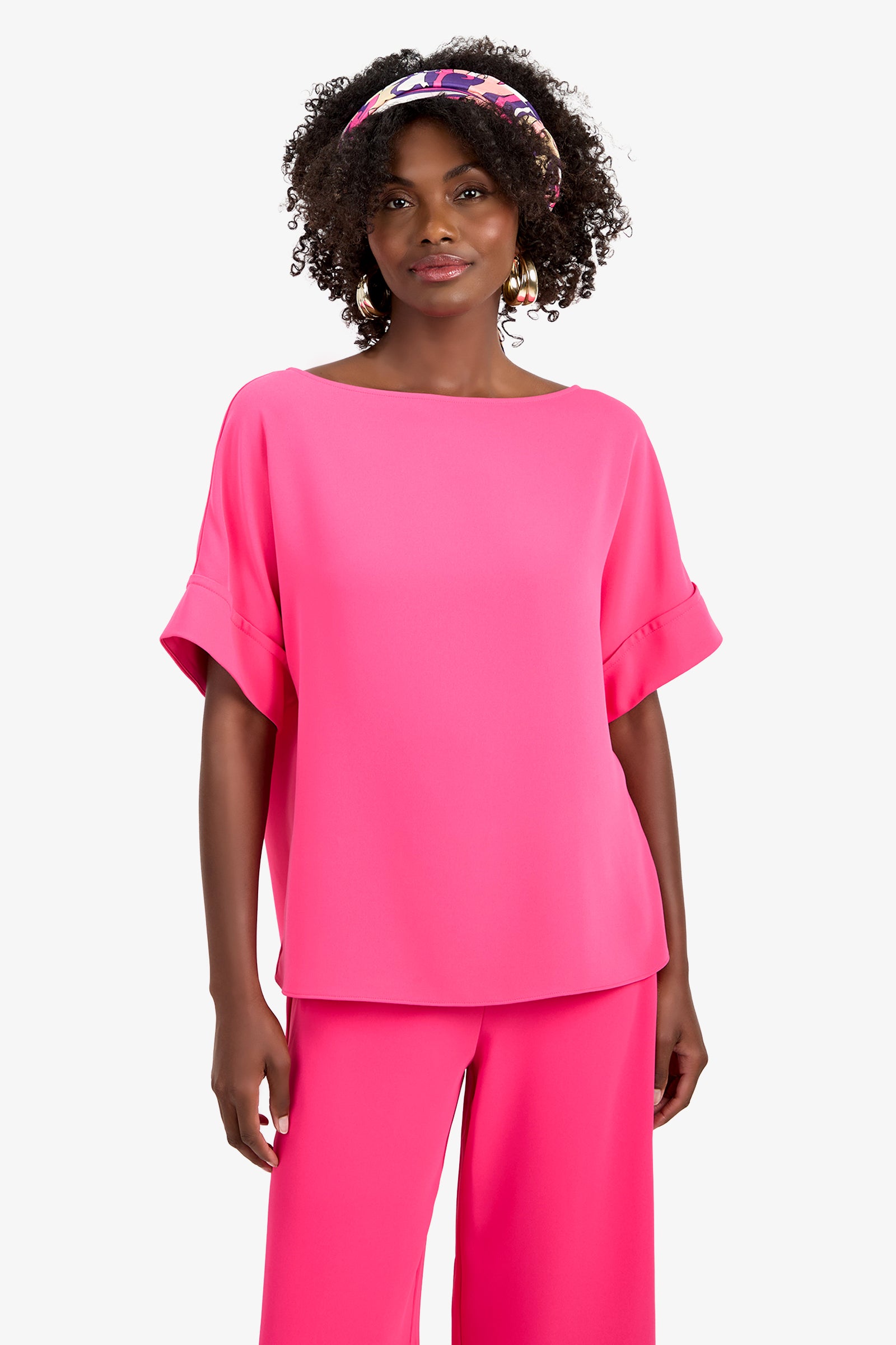 A woman with curly hair and a colorful headband models the SOFIE TOP, a bright pink loose-fit polyester top with cuffed short sleeves, paired with matching wide-leg pants, standing against a plain white background.