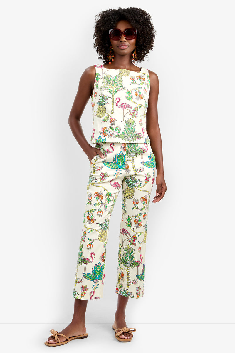 A woman with curly hair, large round sunglasses, and sandals wears the Venya Top—a sleeveless, white tropical-printed top paired with matching flamingo and plant print pants. She stands confidently with one hand in her pocket.