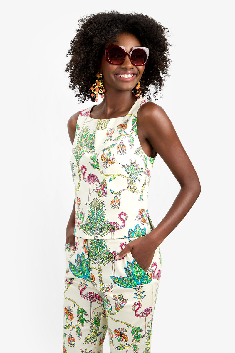 A woman with curly hair smiles, wearing large sunglasses, colorful earrings, and the VENYA 2 TOP—a sleeveless, white cotton-spandex top with a vibrant tropical flamingo and leaf print. Her hands are in her pockets.