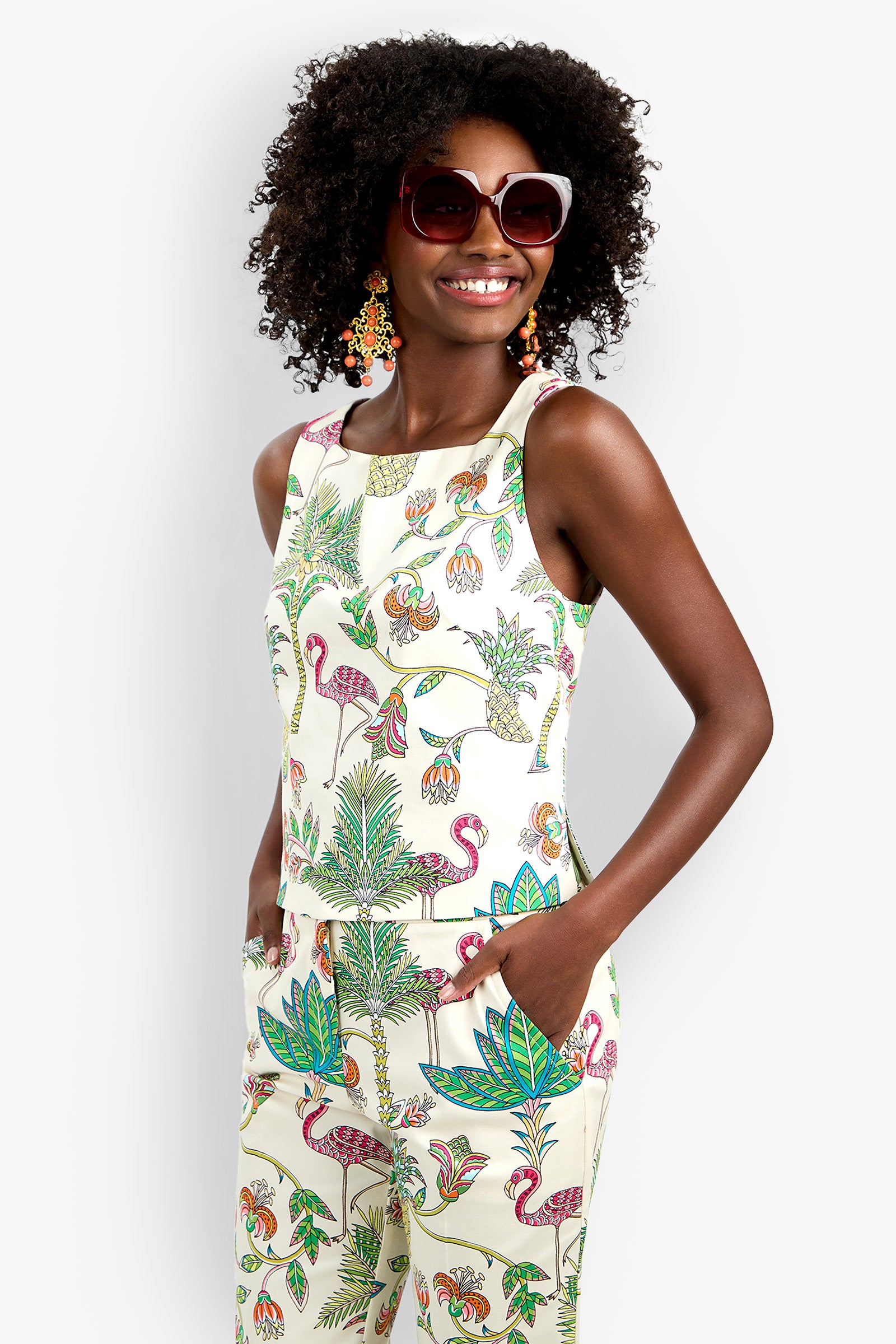 A woman with curly hair smiles, hands in pockets, wearing the Venya Top and matching pants—a sleeveless set with a vibrant tropical print featuring flamingos, leaves, and flowers—paired with large sunglasses and colorful earrings.