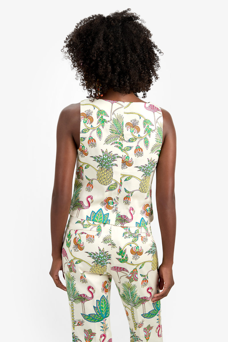 A woman with curly hair is shown from the back wearing the sleeveless, cream-colored VENYA 2 TOP and matching spandex pants, both featuring a colorful tropical print of pineapples, flamingos, monkeys, and leaves against a white background.