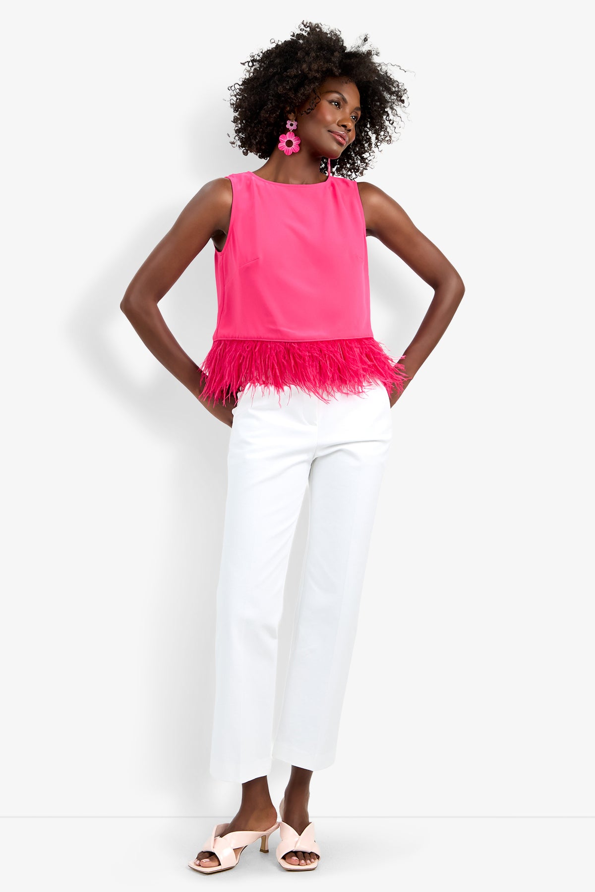 A woman poses against a white background wearing the sleeveless hot pink Rubia Top with feather trim, white wide-leg pants, pink tasseled earrings, and light pink sandals.