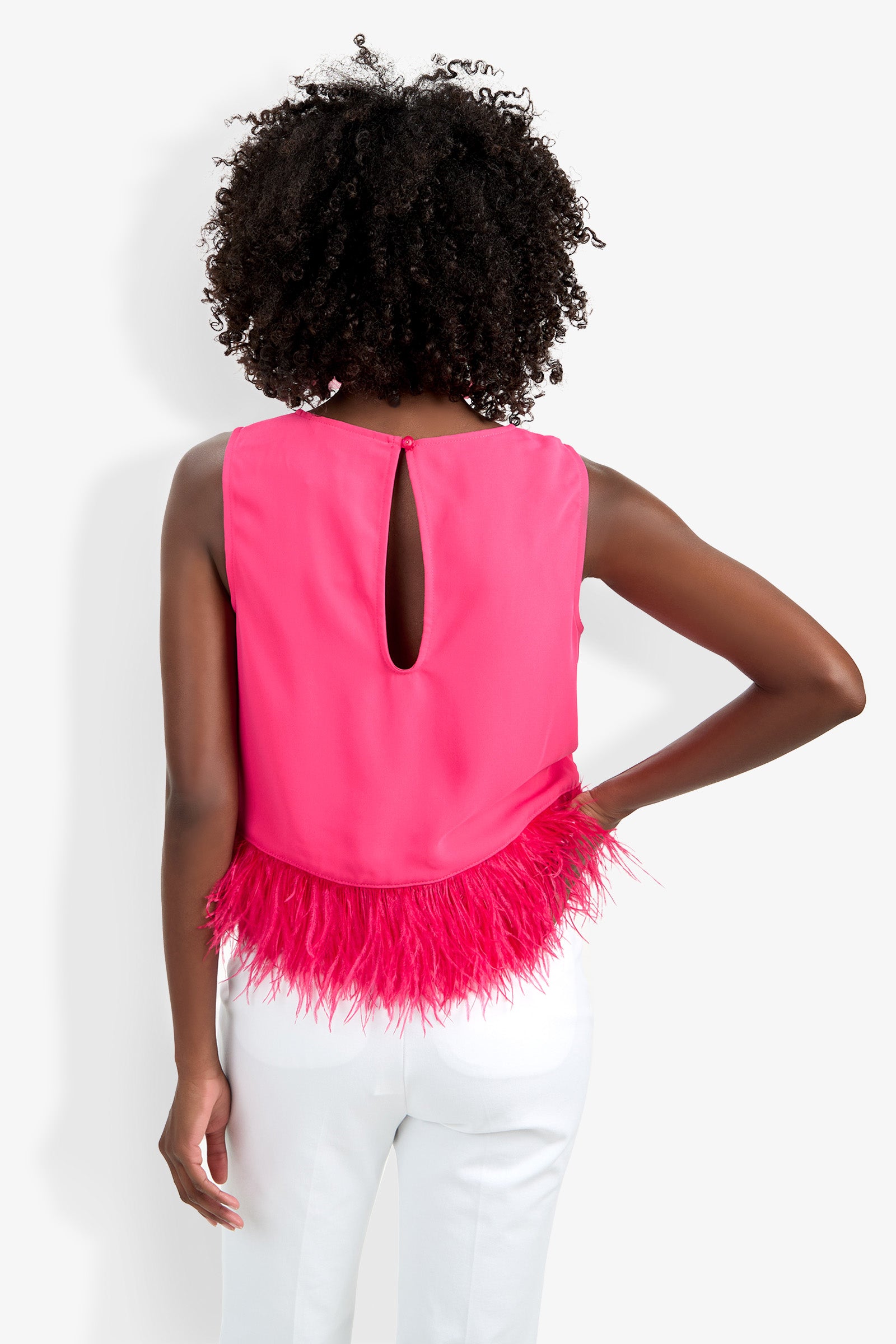 A woman with curly hair stands facing away, wearing the sleeveless Rubia Top in hot pink with a keyhole back and feather trim at the hem, paired with white pants. One arm is bent at the elbow.