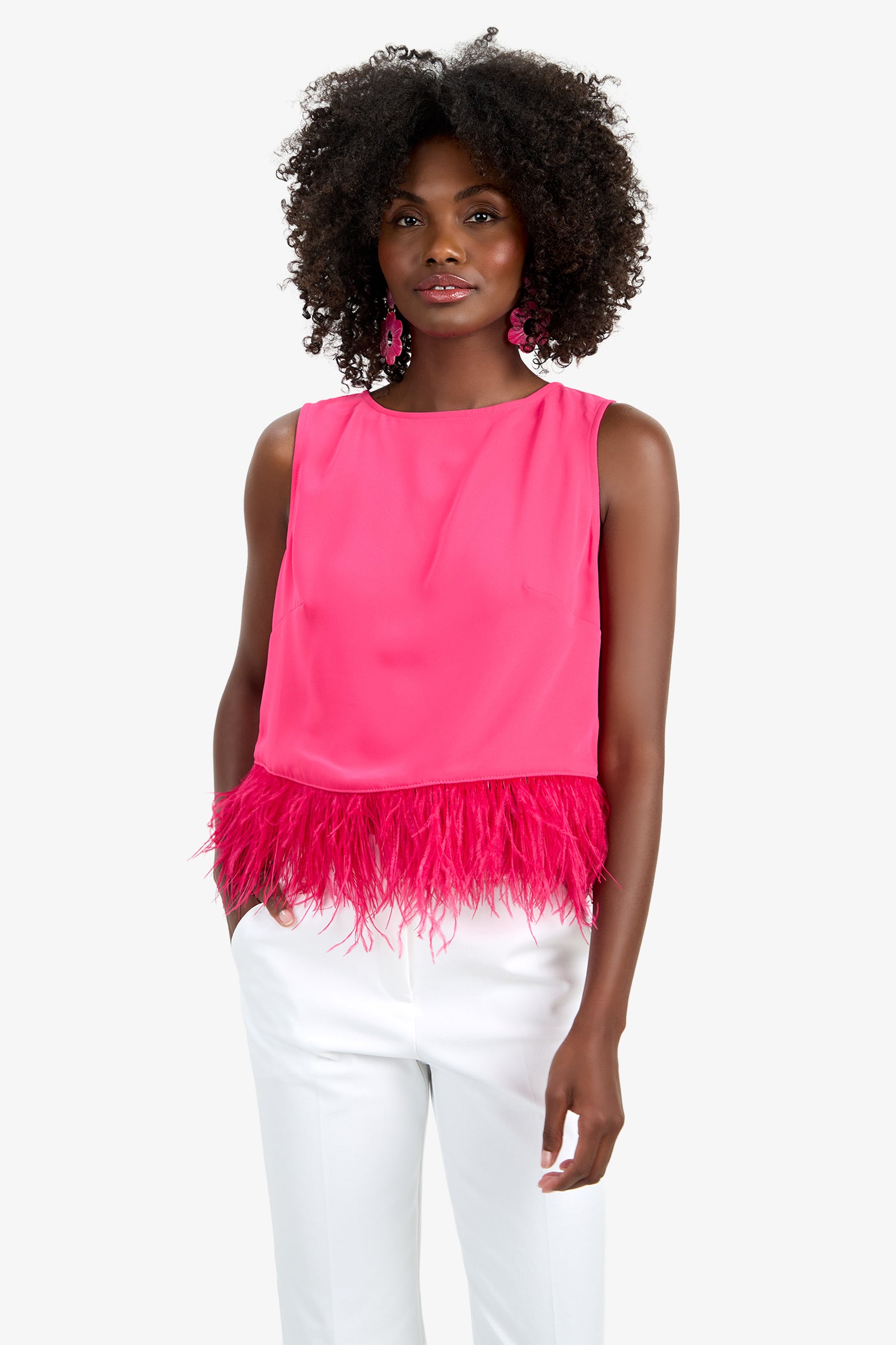 A woman with curly hair and pink earrings stands against a plain white background wearing the Rubia Top—a sleeveless, bright pink top with feather trim at the hem—paired with white pants.