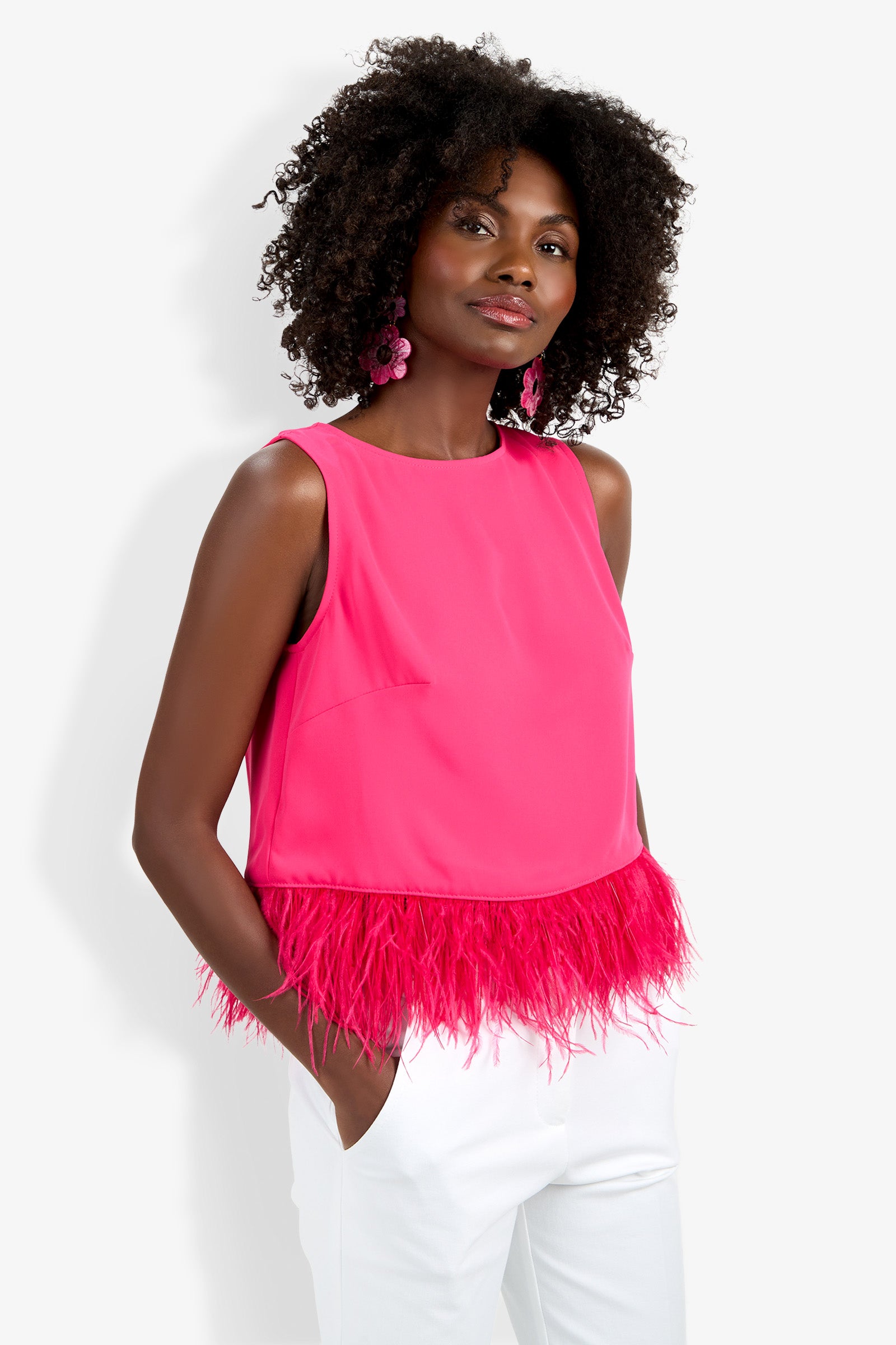 A woman stands against a plain white background, wearing the Rubia Top—a bright pink sleeveless top with feather trim at the hem—paired with white pants and large pink earrings, her hands tucked in her pockets.