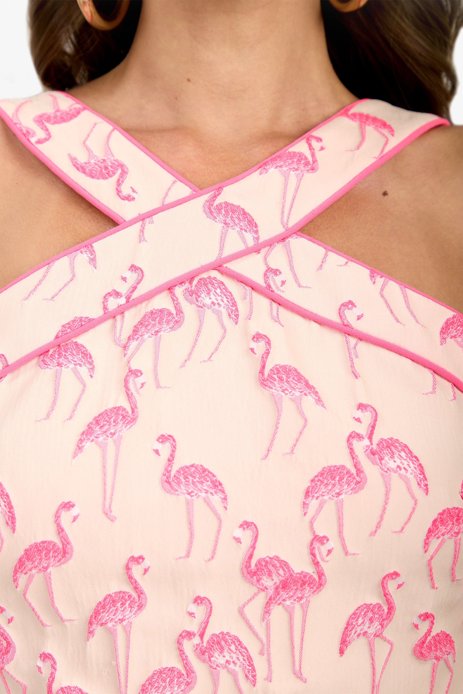 Close-up of the Piped Wanona Top, a light pink garment with crisscrossed straps and an embroidered pink flamingo pattern, highlighting the upper chest and neckline area.
